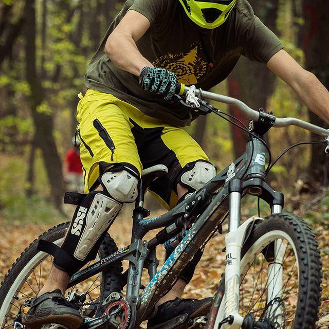 A mountain biker wearing a bright yellow helmet, a green t-shirt, and knee pads, riding a mountain bike on a trail covered with autumn leaves surrounded by trees. Bike Park Josutnjak mountain bike trail.