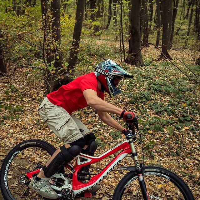 A cyclist wearing a helmet and protective gear rides a red mountain bike on a trail surrounded by trees and autumn foliage. Bike Park Josutnjak mountain bike trail.