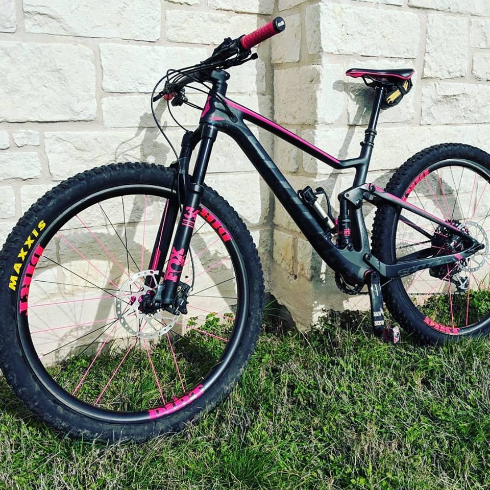 Scott Spark 910: A black and pink mountain bike with large tires, leaning against a stone wall. The bike features a front suspension fork and a sleek frame design, with details such as a pink handlebar grip and branding on the wheels. The grass is visible in the foreground.