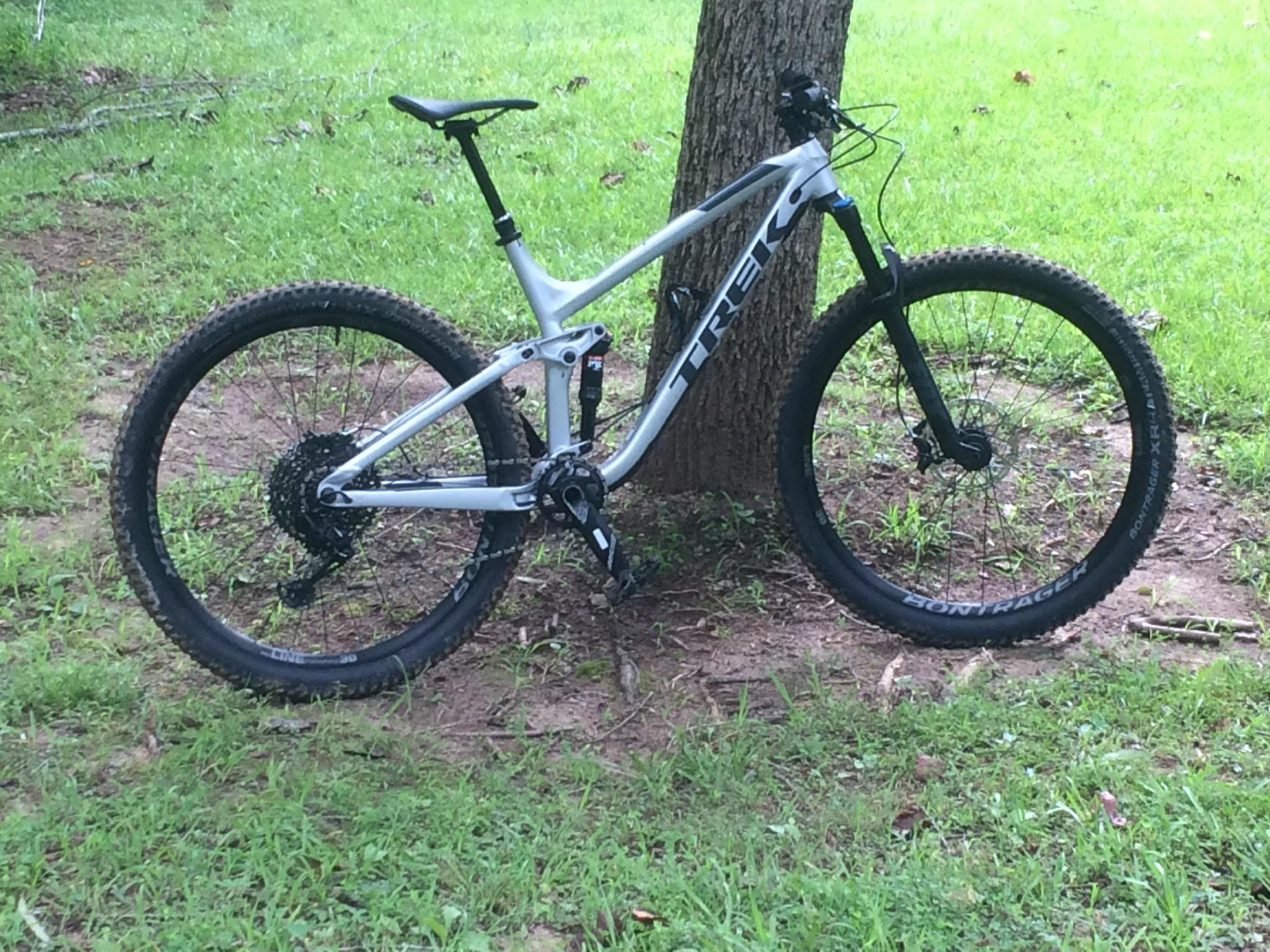 Trek Fuel EX 8: A silver Trek mountain bike leaning against a tree, positioned on a grassy area, with a dirt path visible in the background. The bike features wide tires and a suspension system, indicating its suitability for off-road cycling.