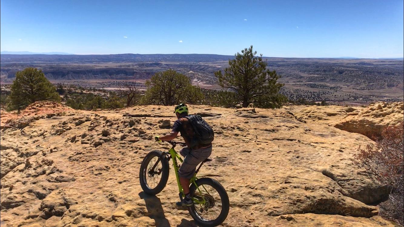 A mountain biker navigating rocky terrain with a scenic view of valleys and distant mountains under a clear blue sky. The rider is wearing a green helmet and a backpack, positioned on a large rock surface surrounded by sparse vegetation. Red Mesa mountain bike trail.