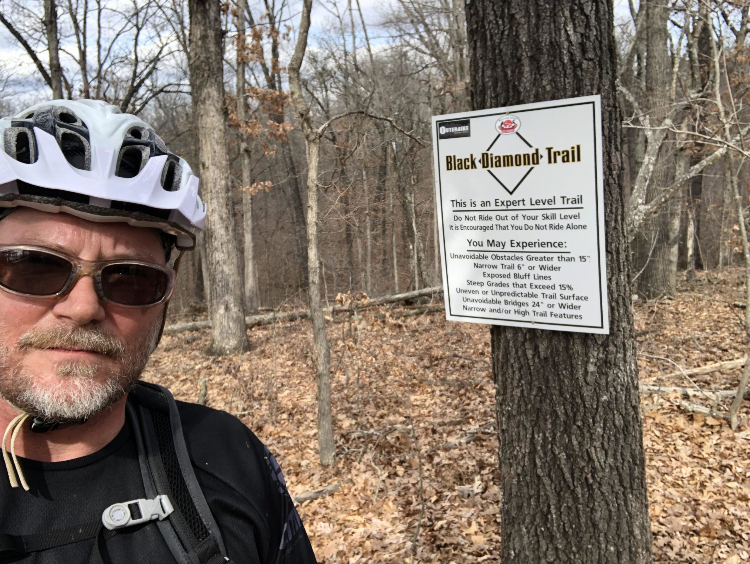A person wearing a helmet and sunglasses stands in front of a trail sign indicating the "Black Diamond Trail," which is marked as an expert level biking trail. The sign warns of potential hazards such as steep grades, narrow paths, and unavoidable obstacles. The background features a wooded area with bare trees and fallen leaves. Back 40 mountain bike trail.