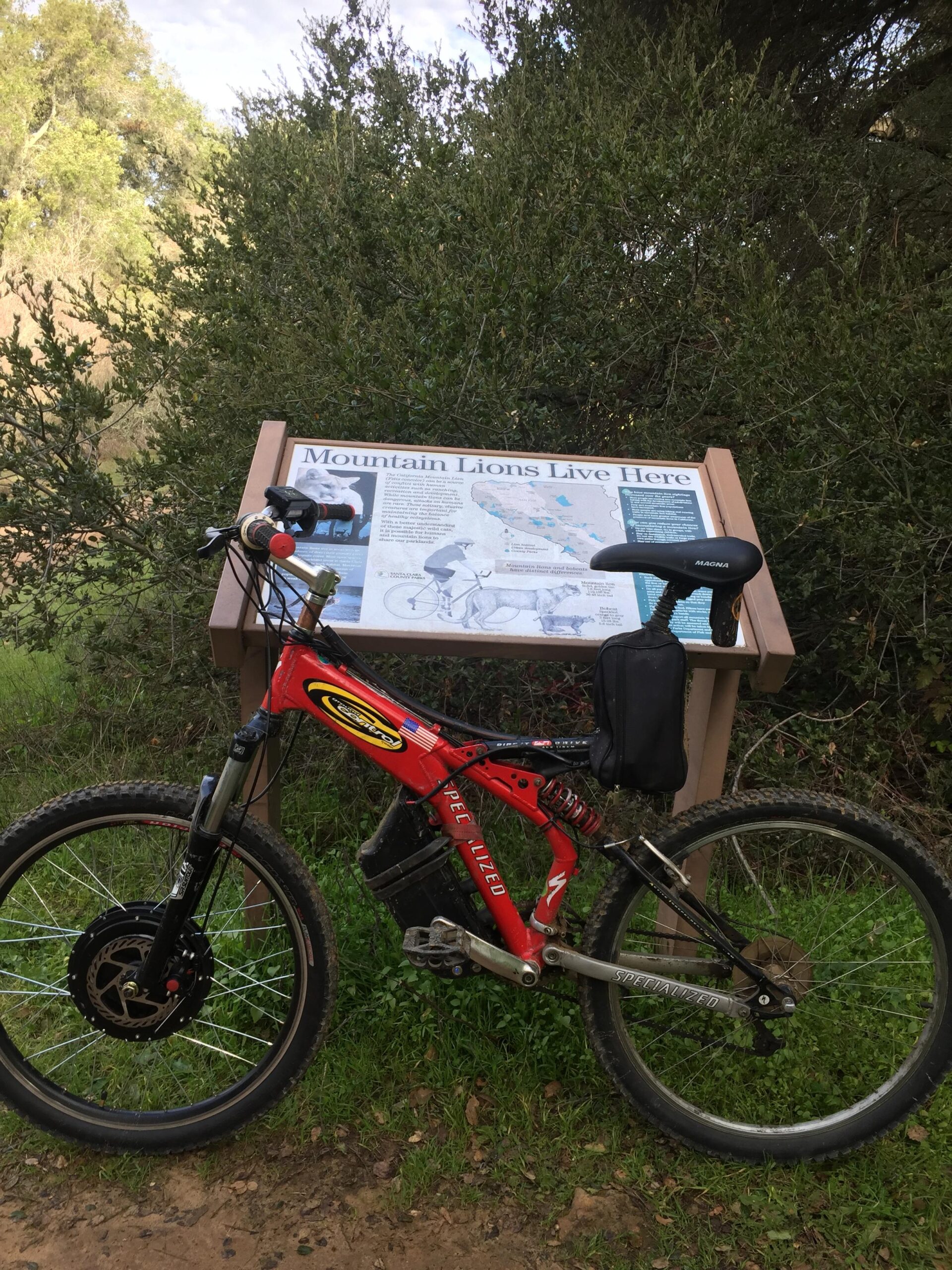 Specialized Ground control: A red mountain bike parked near a wooden informational sign that reads "Mountain Lions Live Here," surrounded by greenery in a natural outdoor setting. The sign features images and text about mountain lions and their habitat.