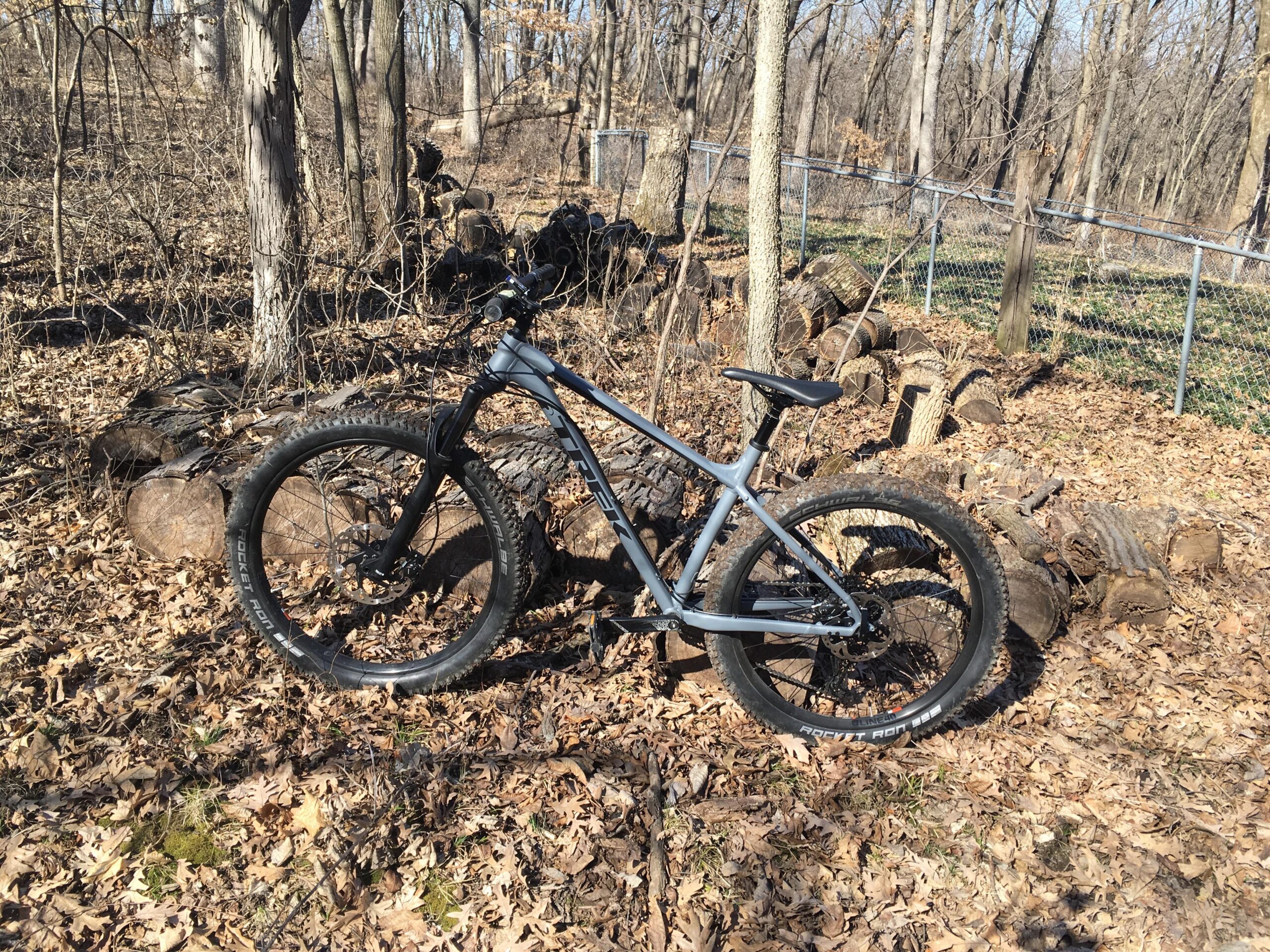 Trek Roscoe 8: A silver mountain bike leaning against a stack of logs in a forested area, surrounded by fallen leaves and trees.