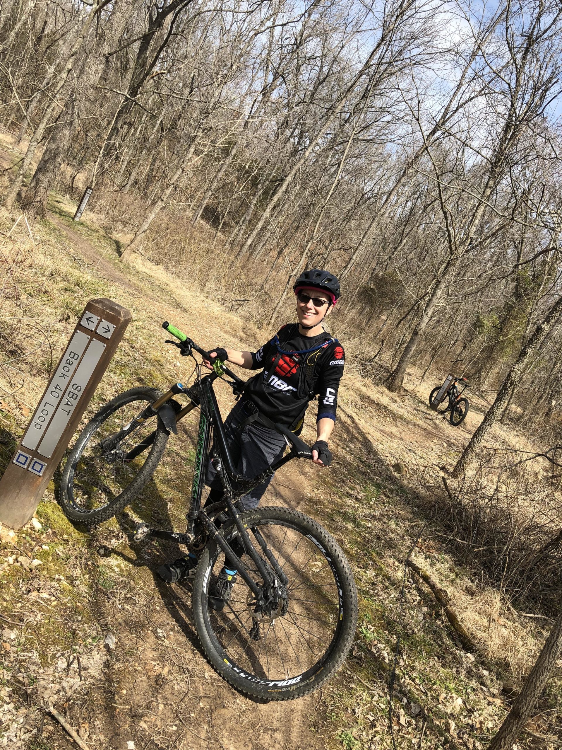 A mountain biker stands next to a trail sign in a wooded area, showing a well-maintained dirt path surrounded by bare trees. The biker is smiling and wearing a helmet and protective gear, while holding the handlebars of a black mountain bike. Another biker is visible in the background on the same trail. Back 40 mountain bike trail.