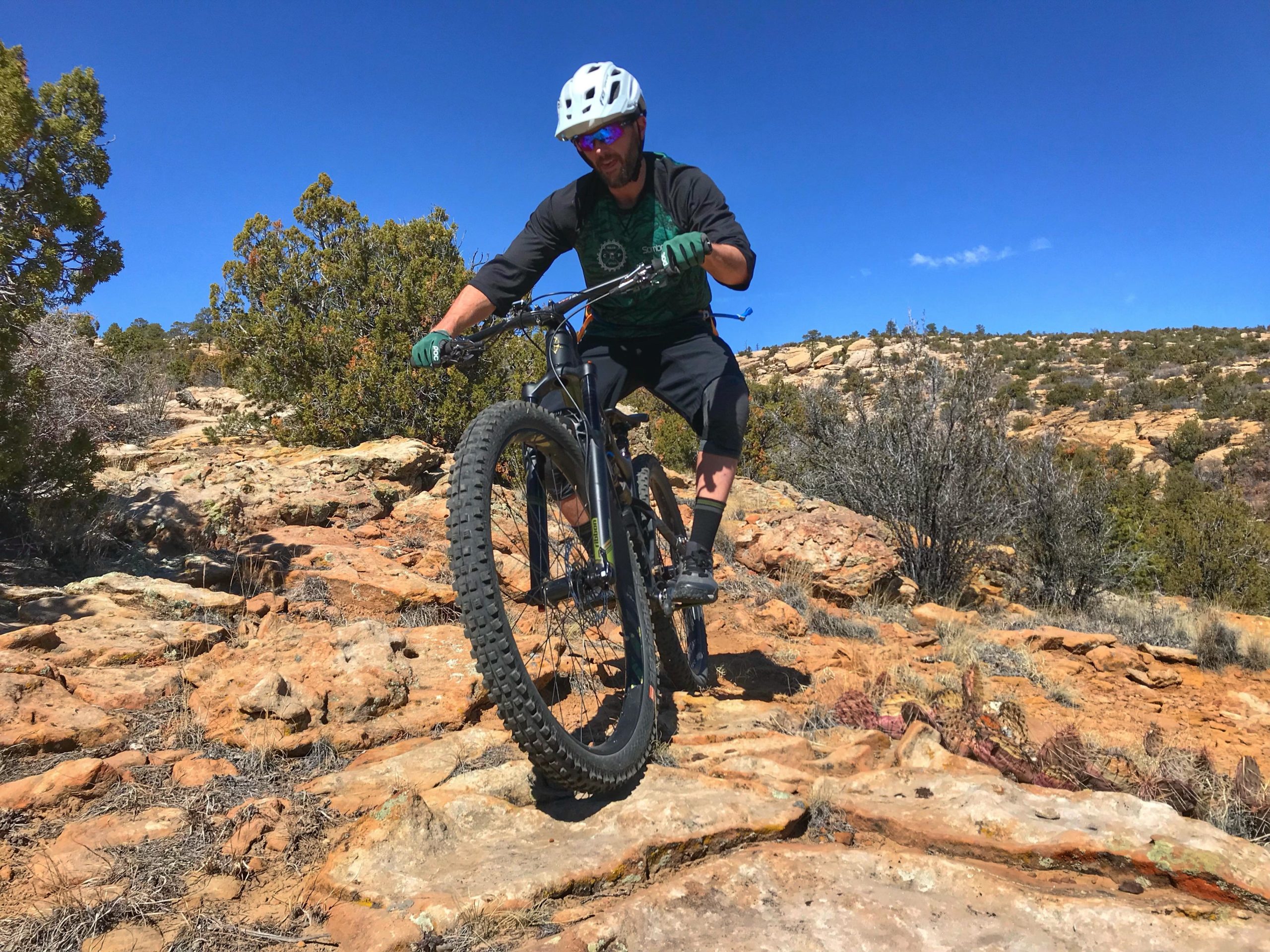 A mountain biker navigating a rocky trail on a bright, sunny day. The cyclist is wearing a helmet and sunglasses, and is skillfully maneuvering over uneven terrain amidst sparse vegetation and rocky outcrops. The landscape features a clear blue sky in the background. Red Mesa mountain bike trail.