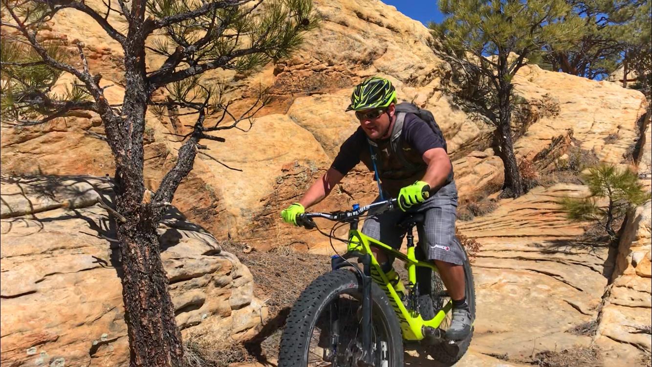 A mountain biker navigates a rocky trail surrounded by natural vegetation, wearing a helmet and gloves. The bike features large tires, and the rider is focused on the path ahead. The terrain is dry with sunlit rock formations in the background. Red Mesa mountain bike trail.
