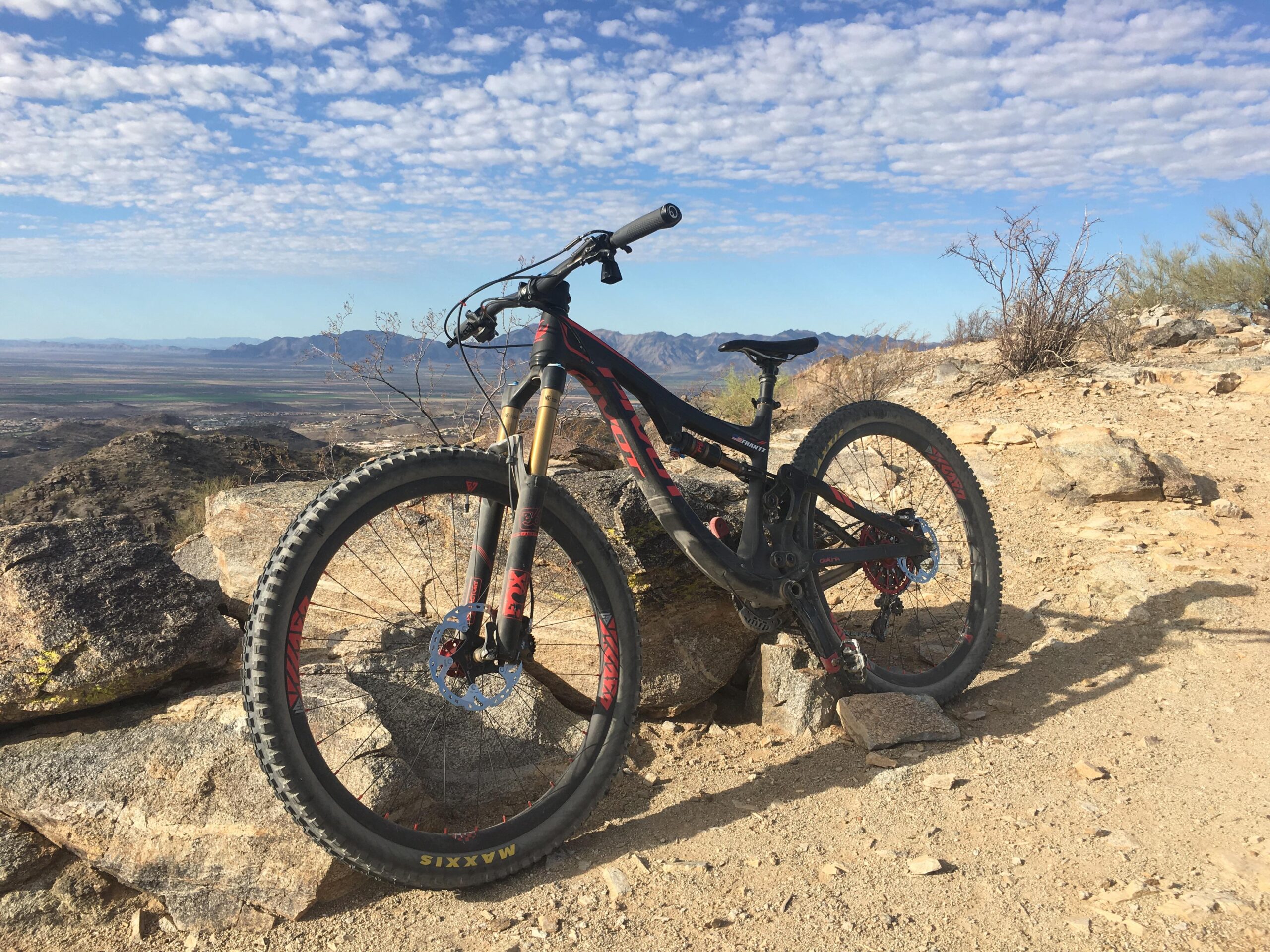 Pivot SwitchBlade: A mountain bike resting on a rocky surface, with a scenic view of a valley and distant mountains under a partly cloudy sky. The bike features prominent tires and suspension, positioned against a backdrop of rugged terrain.