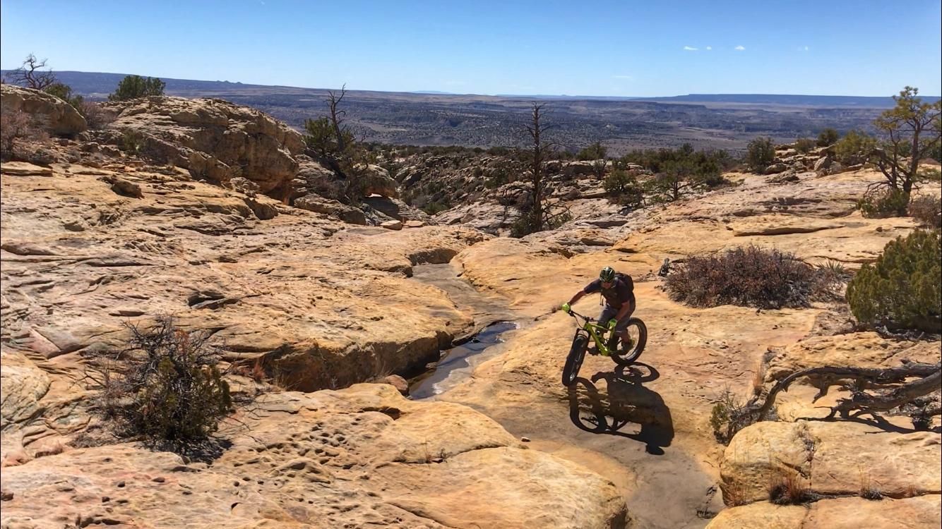 A mountain biker navigates a rocky terrain under a clear blue sky, surrounded by desert landscape and sparse vegetation. The terrain features large, rough boulders and a narrow channel of water running through the rocks. Red Mesa mountain bike trail.
