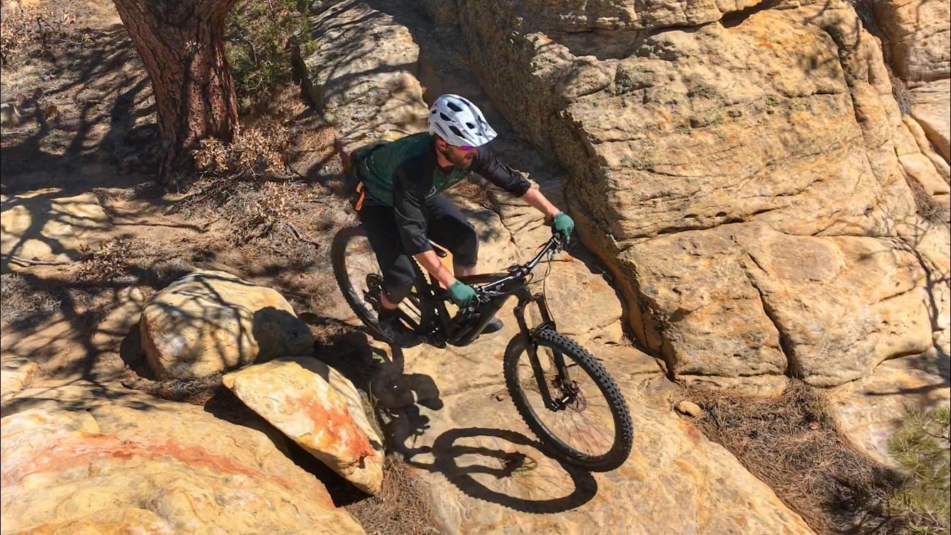 A mountain biker navigating rocky terrain on a sunny day. The cyclist is wearing a helmet and gloves, showcasing skills as they maneuver over large boulders and uneven ground. Surrounding vegetation adds to the outdoor atmosphere, highlighting an adventurous biking experience. Red Mesa mountain bike trail.