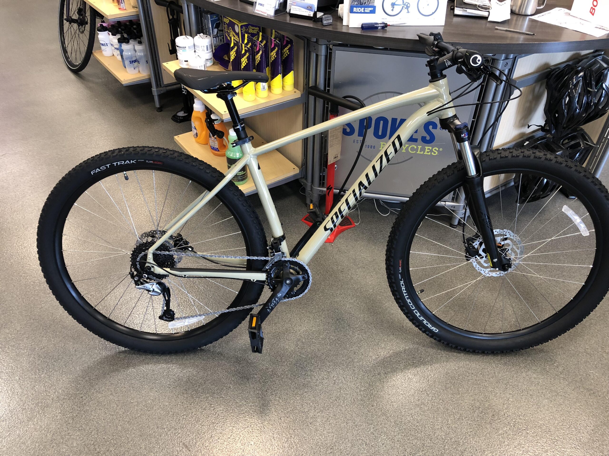 Specialized Rockhopper Comp 29: Alt text: A specialized mountain bike with a light-colored frame, front suspension, and thick tires, displayed inside a bicycle shop. Shelves in the background contain bike accessories and water bottles.
