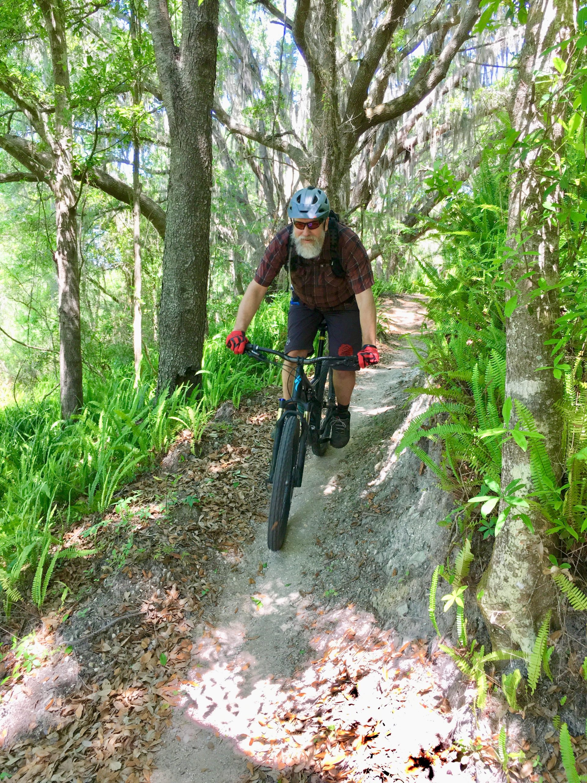 A person riding a mountain bike on a narrow, dirt trail surrounded by lush greenery and trees. The cyclist is wearing a helmet, sunglasses, and gloves, focused on navigating the path. Sunlight filters through the trees, creating dappled light on the ground. Balm Boyette Scrub Preserve mountain bike trail.