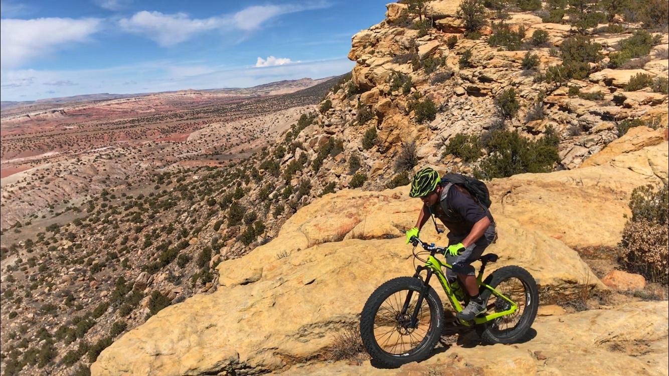 A person riding a fat tire mountain bike on rocky terrain with a vast desert landscape in the background. The rider is wearing a helmet and gloves, and appears focused as they navigate the challenging path. The sky is bright with some clouds, highlighting the rich colors of the surrounding land. Red Mesa mountain bike trail.