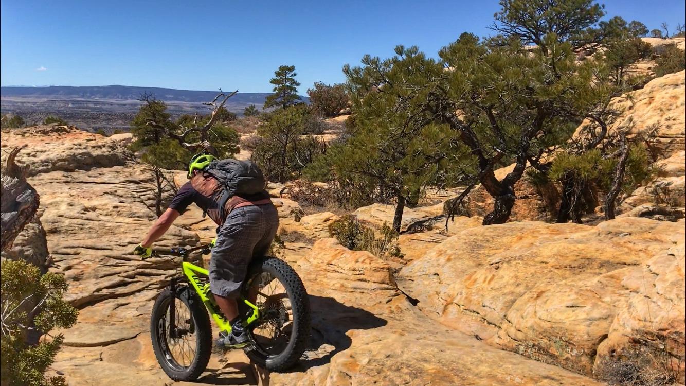 A person riding a mountain bike on rocky terrain in a natural outdoor setting, surrounded by trees and distant mountains under a clear blue sky. The cyclist is wearing a helmet and carrying a backpack. Red Mesa mountain bike trail.