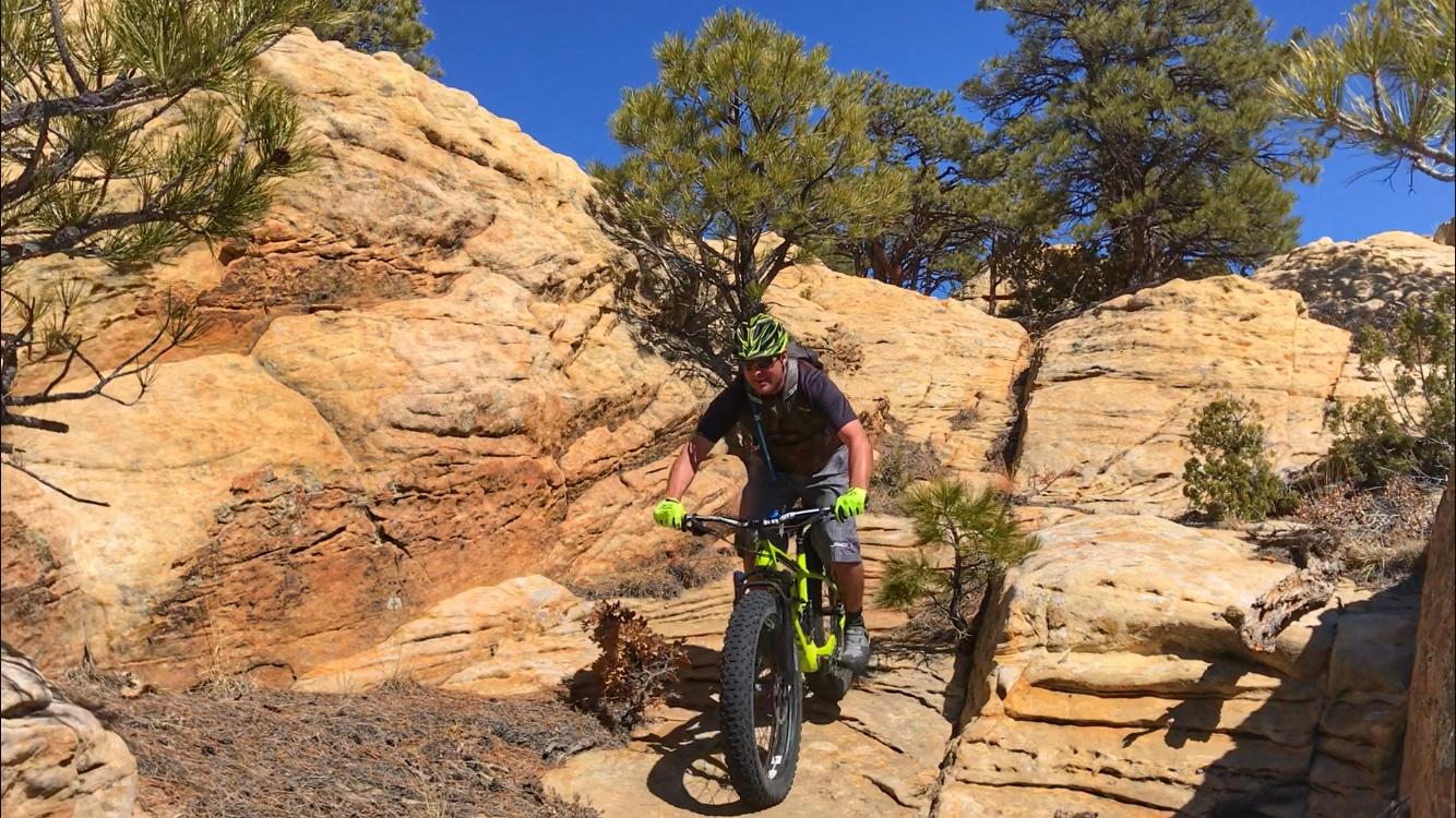 A person riding a fat bike on a rocky trail surrounded by trees and clear blue skies. The cyclist is wearing a helmet and green gloves, focused on navigating the terrain. Red Mesa mountain bike trail.