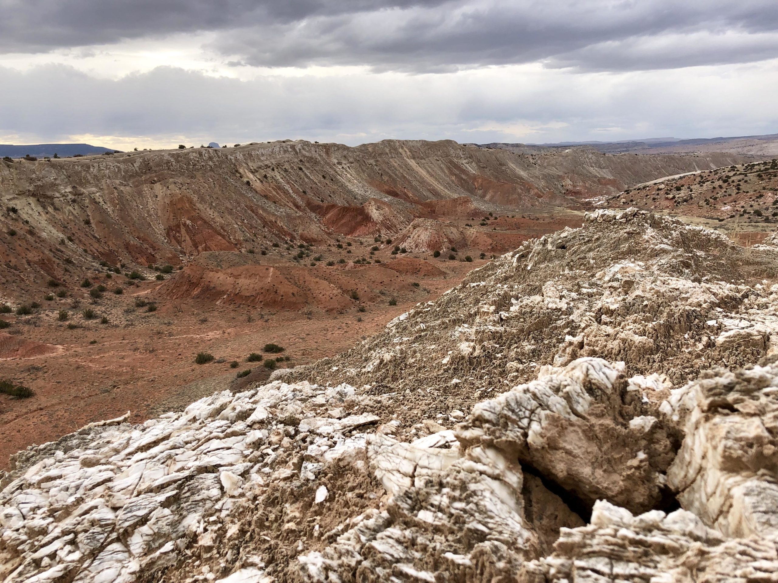 A rocky landscape featuring rugged hills with layers of red and brown earth under a cloudy sky. In the foreground, there are white, textured rock formations, while the background reveals a series of undulating hills and sparse vegetation. White Ridge Bike Trails mountain bike trail.