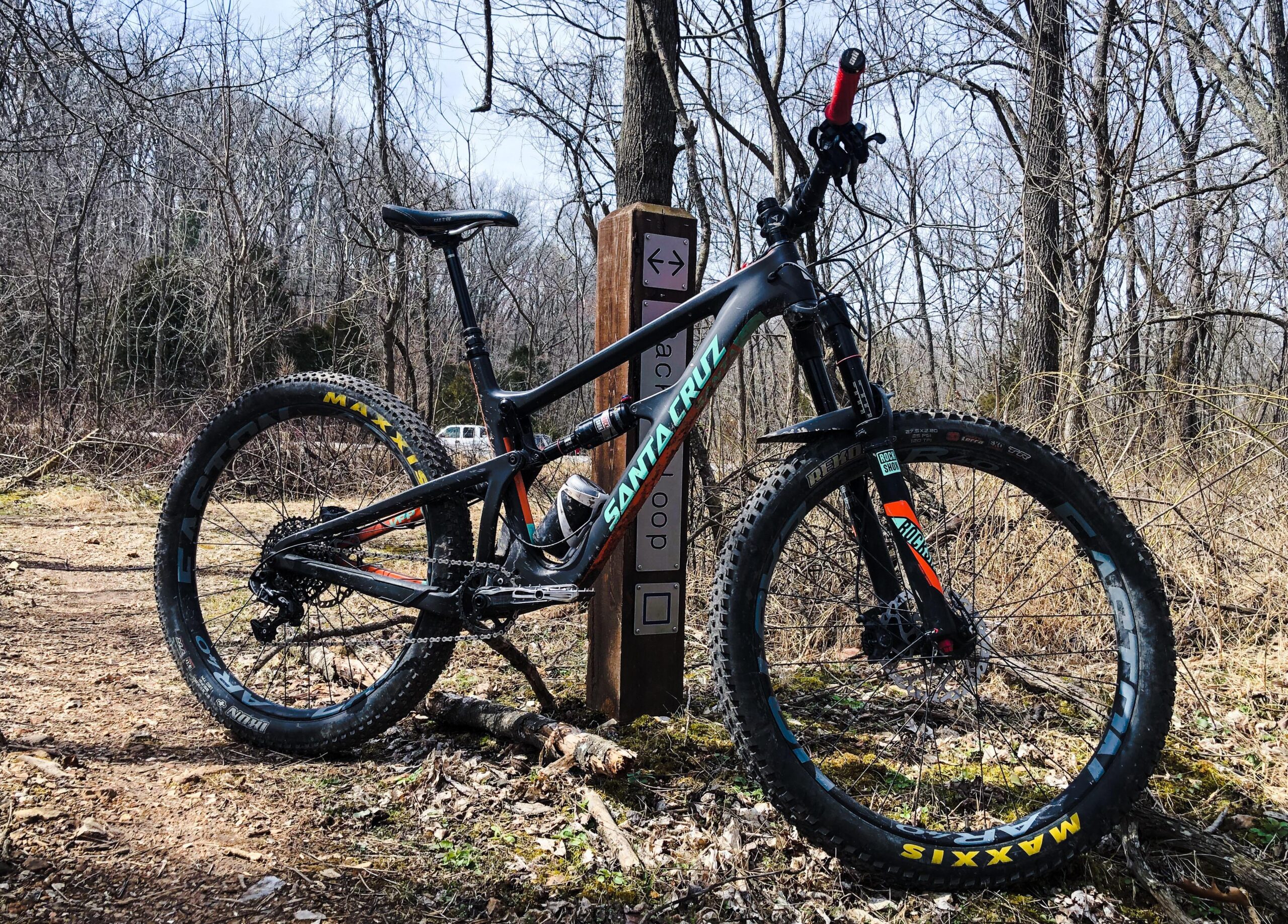 Santa Cruz Hightower LT: A black mountain bike with vibrant decals, leaning against a wooden trail sign in a wooded area. The bike features wide tires with a "Maxxis" logo, and the background includes bare trees and underbrush, suggesting an early spring setting.