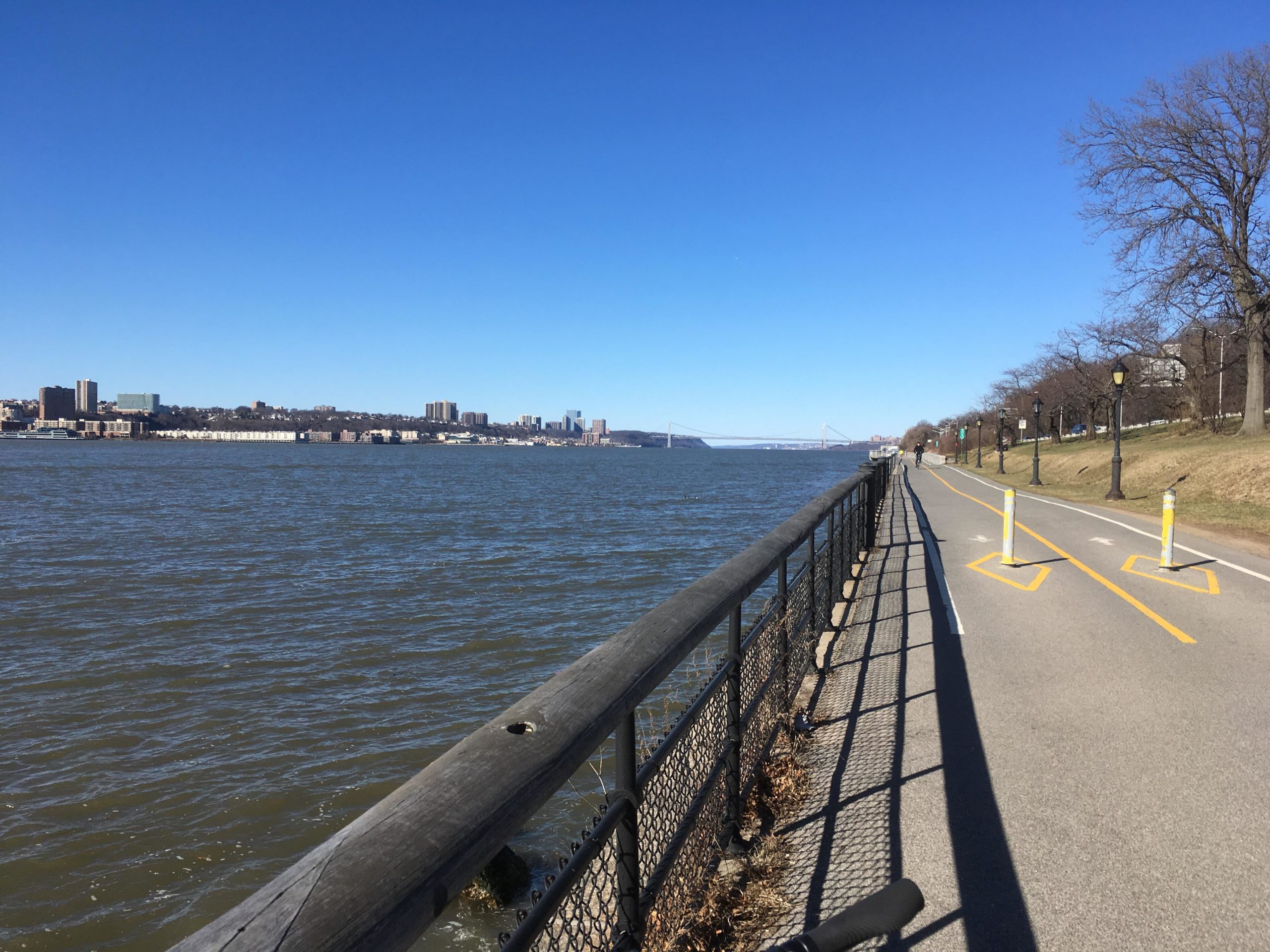 A view of the Hudson River with a paved path along the waterfront. The image features blue skies, a railing along the path, and city buildings in the background. There are trees lining the path and a few people walking or biking. West Street Greenway mountain bike trail.