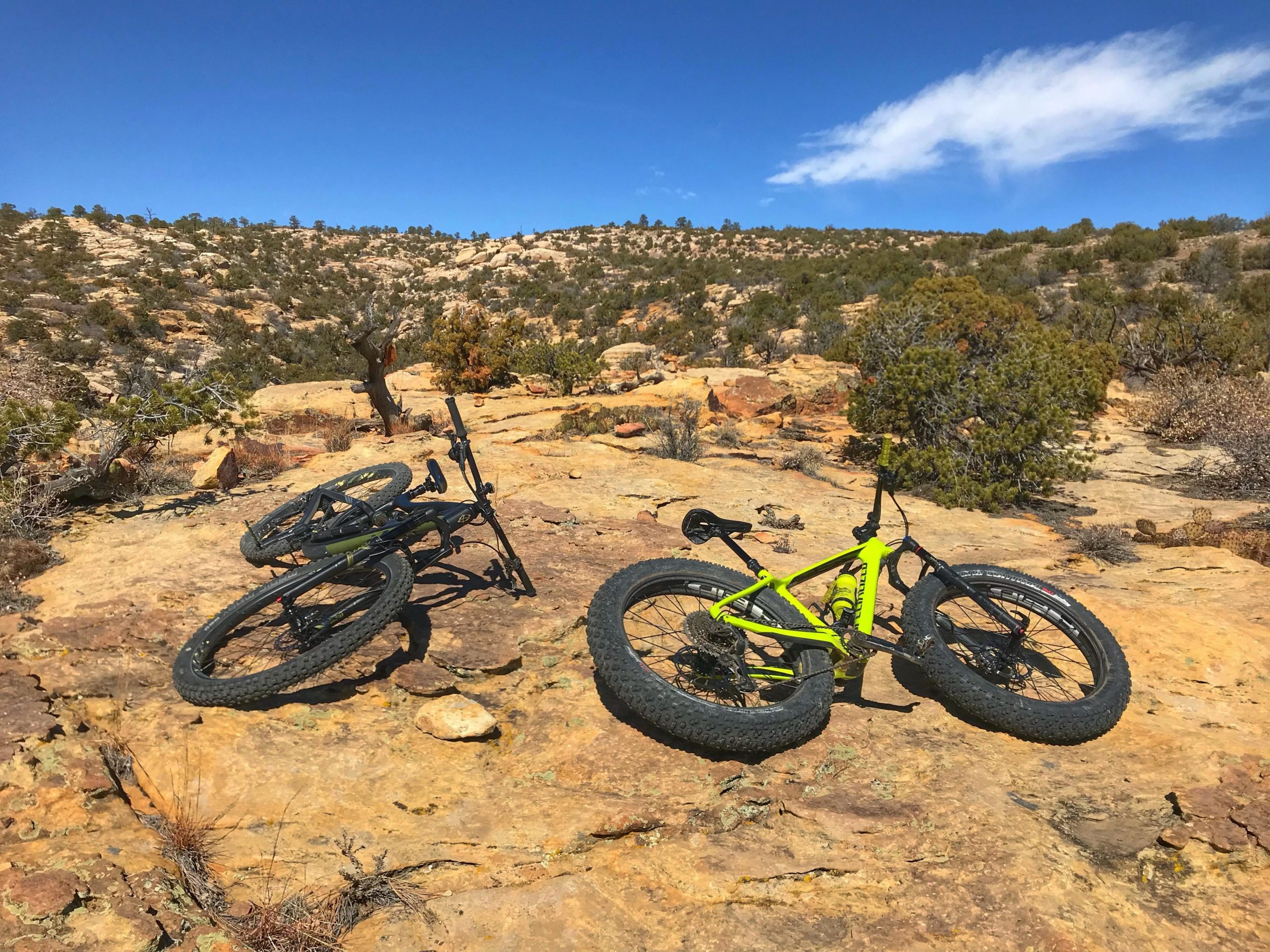 Two mountain bikes are lying on a rocky landscape under a clear blue sky. One bike is bright green, while the other is black. In the background, rugged hills and sparse vegetation can be seen. The scene captures a sunny day, highlighting the natural terrain where mountain biking can occur. Red Mesa mountain bike trail.