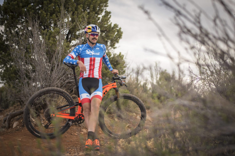A cyclist wearing a colorful jersey featuring the American flag sits on a mountain bike, surrounded by natural vegetation. The bike has large tires and an orange frame, and the cyclist is positioned on a rocky trail with trees in the background. The image captures a sense of adventure and outdoor sports.