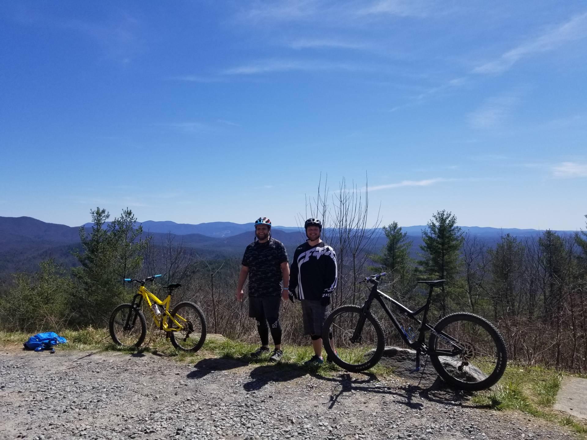 Two mountain bikers standing on a gravel path overlooking a scenic mountain view. One biker is wearing a black and white jersey, while the other is in a dark patterned shirt. They are positioned next to their bikes — a yellow and a black mountain bike — against a backdrop of rolling hills and a clear blue sky. Pinhoti Trail: P1 / Bear Creek Pinhoti mountain bike trail.