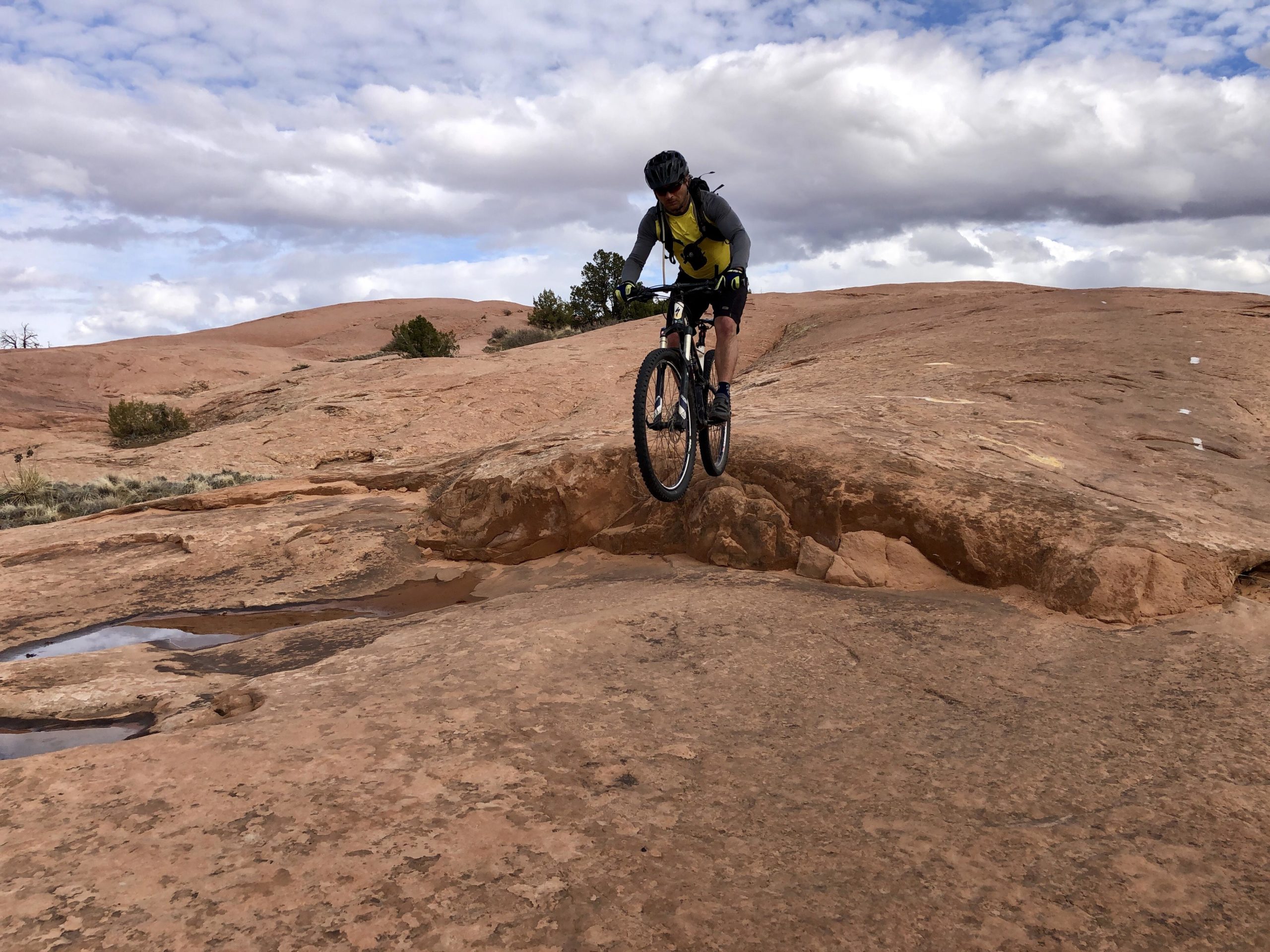 A mountain biker wearing a yellow shirt and gray sleeves jumps over a rocky terrain, with cloudy skies and a distant hill in the background. The scene captures the adventure of biking on rugged trails with patches of water on the rocks. Slickrock mountain bike trail.