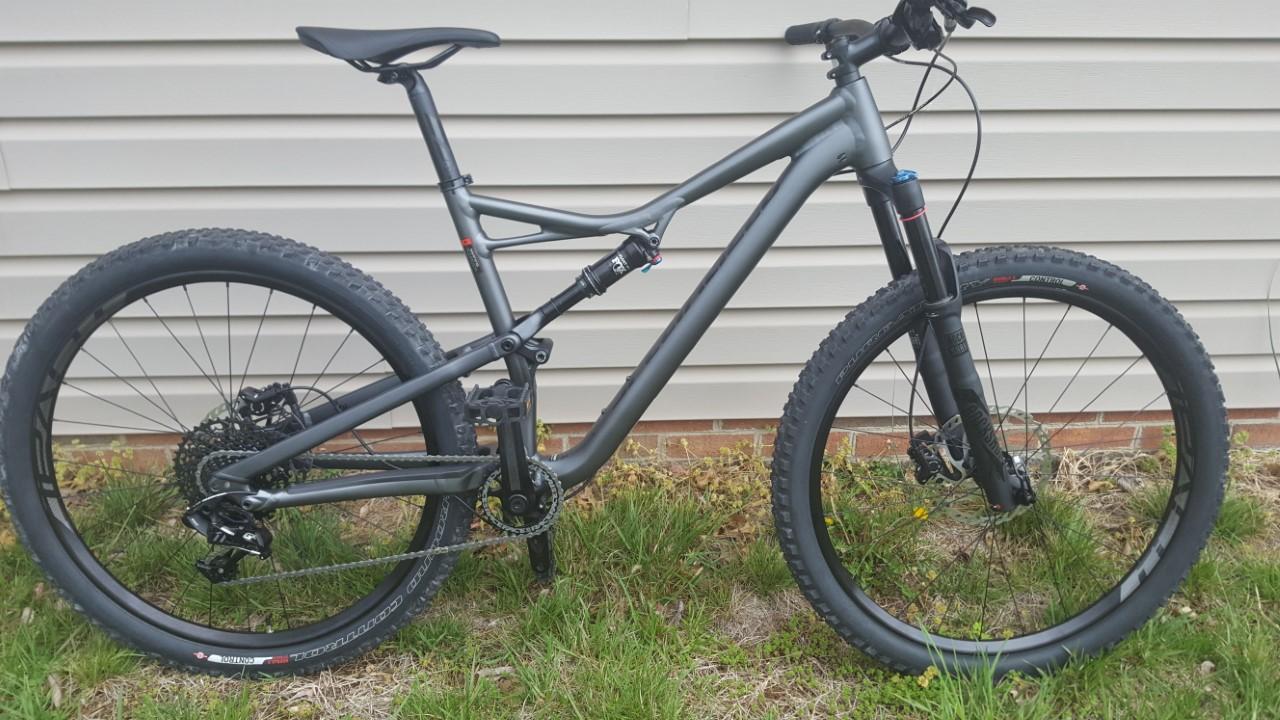 Specialized Camber: Mountain bike with a sleek black frame, front suspension, and wide tires, resting on grass next to a light-colored wall.