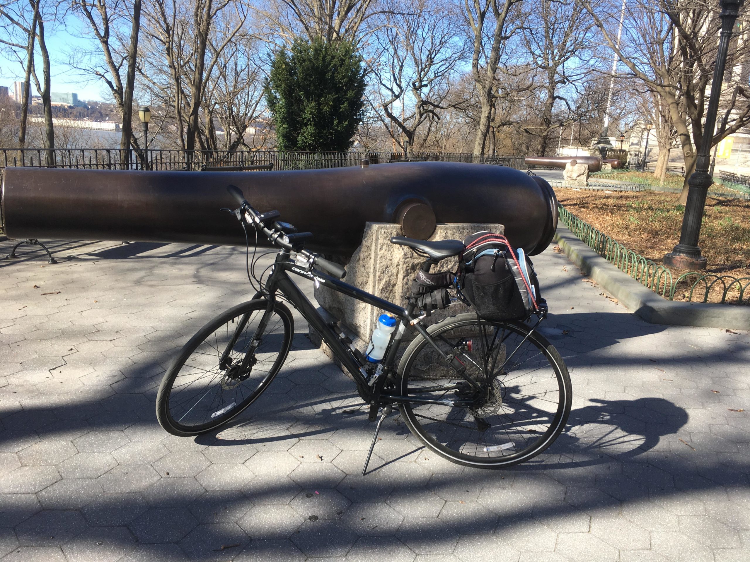 A black bicycle rests on a paved pathway beside a historic cannon. In the background, bare trees and a glimpse of water are visible under a clear blue sky. The area appears to be a park setting with decorative fencing and a lamppost. West Street Greenway mountain bike trail.