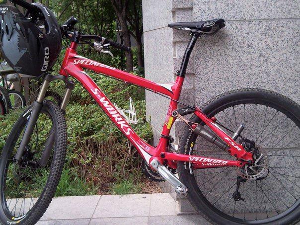 Specialized SWORKS Stumpjumper FSR: A red mountain bike with "S-Works" branding parked against a stone wall, featuring a front suspension fork, knobby tires, and a helmet attached to the handlebars. Green foliage is visible in the background.
