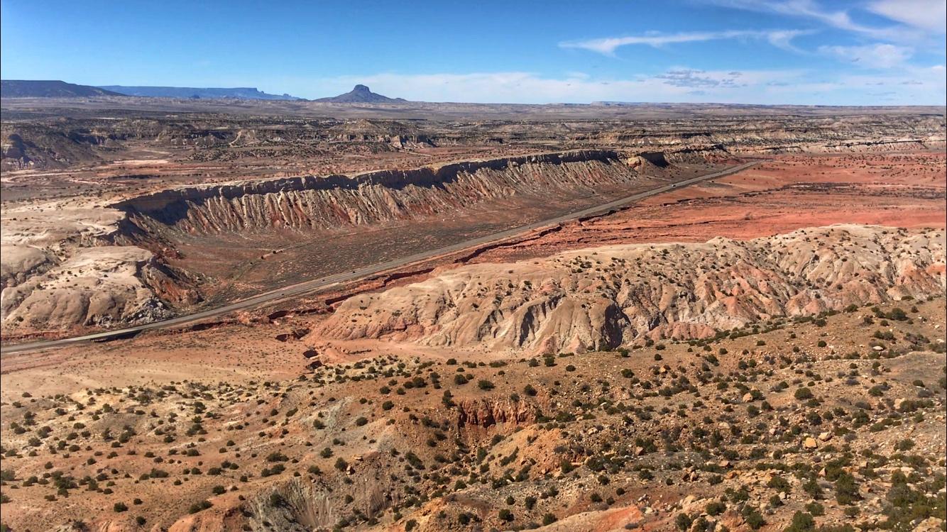 A panoramic view of a rugged desert landscape featuring layered rock formations, a winding road, and scattered vegetation under a clear blue sky. The terrain exhibits various shades of red, brown, and gray, highlighting the natural geological features of the area. Red Mesa mountain bike trail.