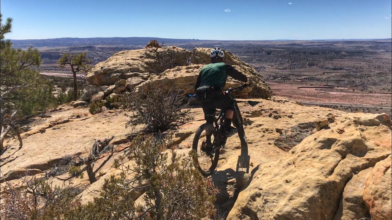 A mountain biker navigating rocky terrain on a cliff edge, with a vast landscape of hills and valleys in the background under a clear blue sky. Red Mesa mountain bike trail.