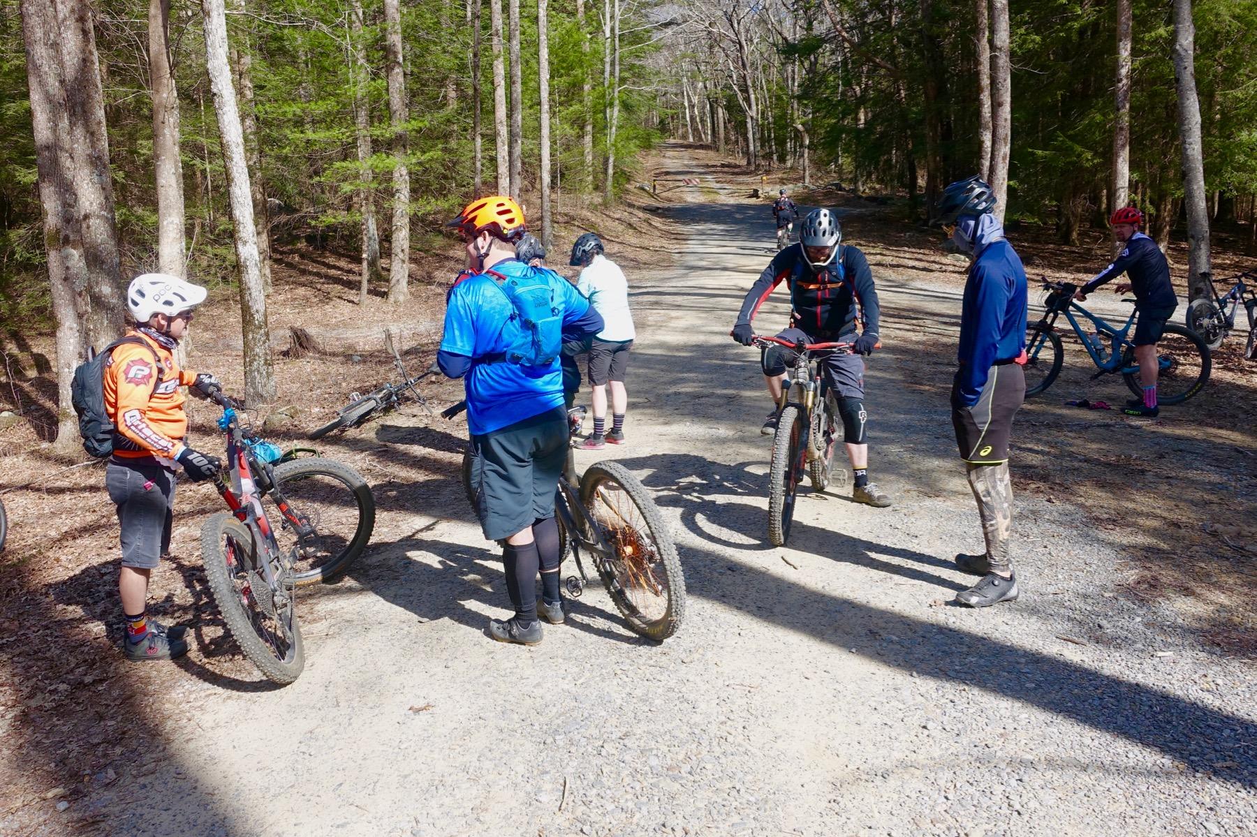 A group of six mountain bikers paused on a gravel trail in a wooded area. They are wearing helmets and biking gear, with some bikes leaned against trees. The scene captures a sunny day with green trees in the background. Windy Gap Trail mountain bike trail.