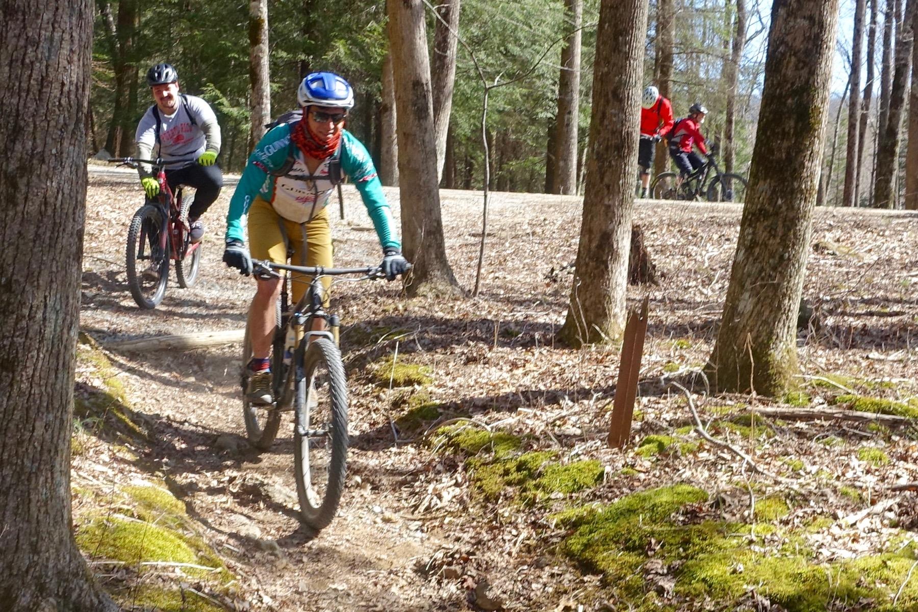 A group of mountain bikers riding on a dirt trail through a sunny forest. The foreground shows a rider in a colorful jersey and shorts, navigating the path, while another rider follows behind. The background features additional bikers and trees, with patches of moss and fallen leaves on the ground. Windy Gap Trail mountain bike trail.