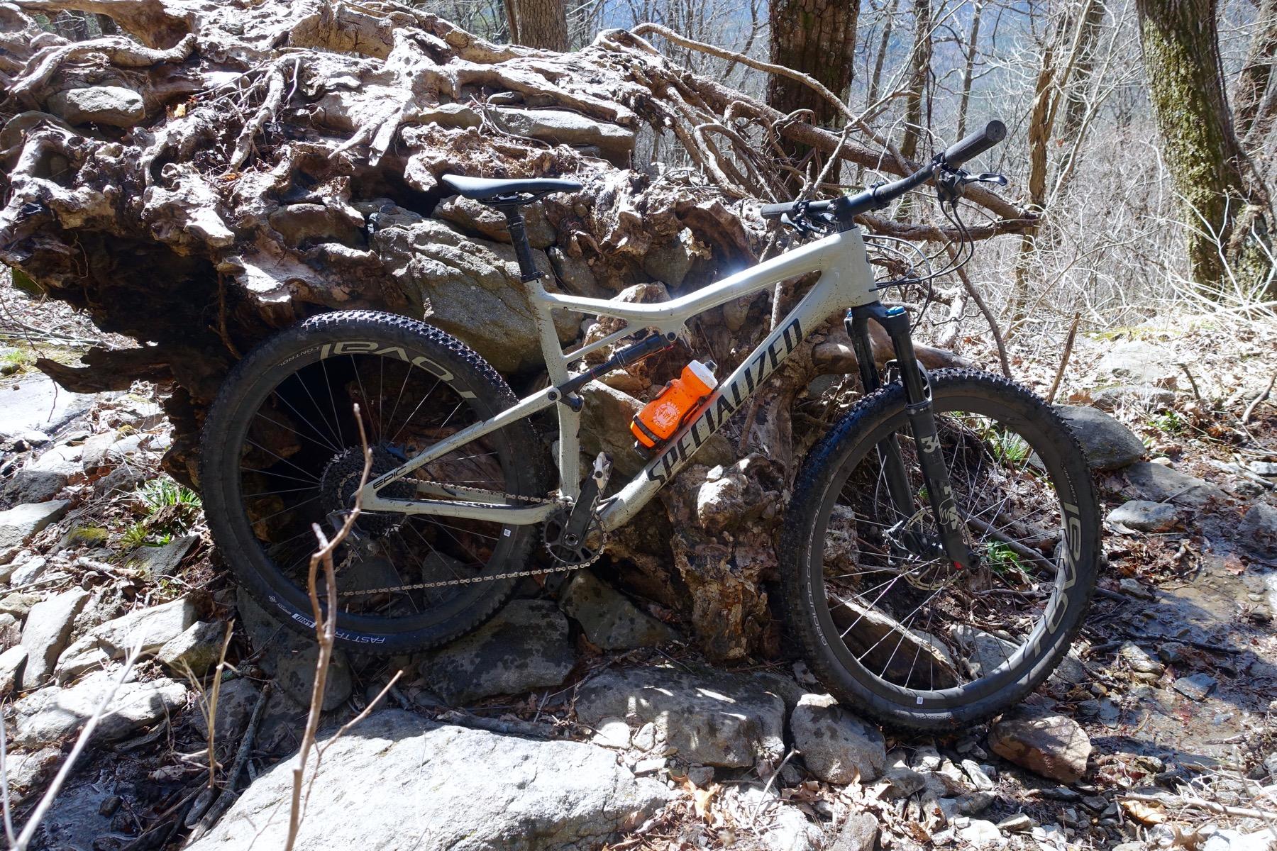 A mountain bike leaning against a large, gnarled tree stump in a forested area. The bike has a gray frame and features a water bottle attached to its frame. Surrounding the bike are rocky terrain and scattered leaves, indicating a natural outdoor setting. Windy Gap Trail mountain bike trail.