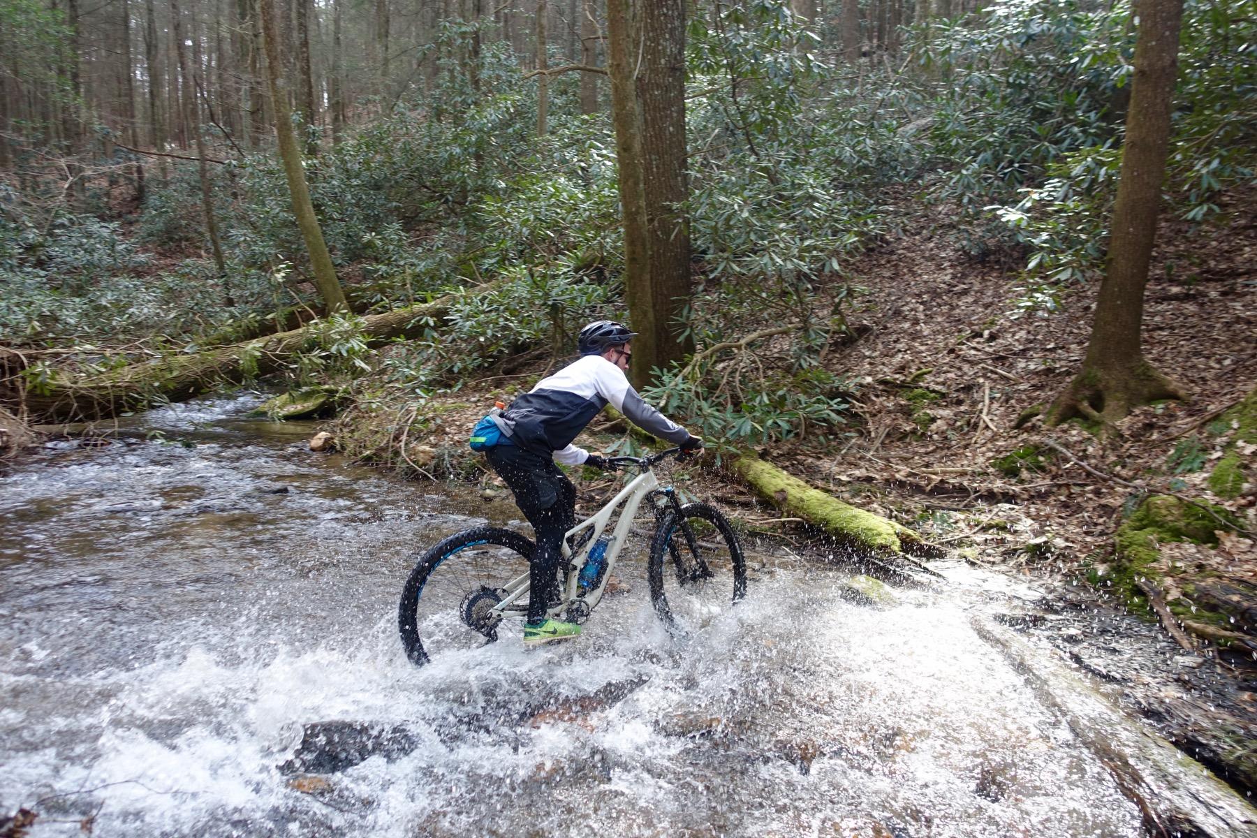 A mountain biker riding through a shallow stream in a forested area, splashing water as he navigates over rocks. The surroundings include trees and lush green foliage, creating a scenic outdoor environment. Pinhoti Trail: Mountaintown Creek Segment mountain bike trail.