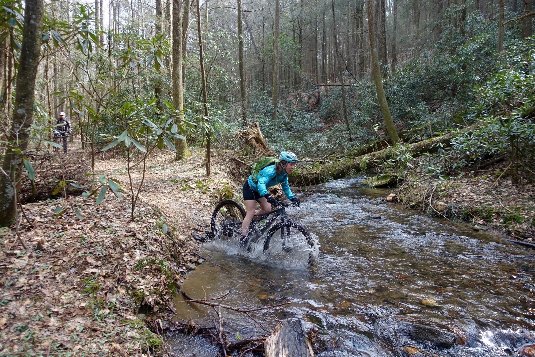 A mountain biker navigating through a shallow stream in a forested area, surrounded by tall trees and lush greenery. The cyclist is splashing through the water, wearing a helmet and a brightly colored jacket, while another person can be seen in the background on the path. Pinhoti Trail: Mountaintown Creek Segment mountain bike trail.