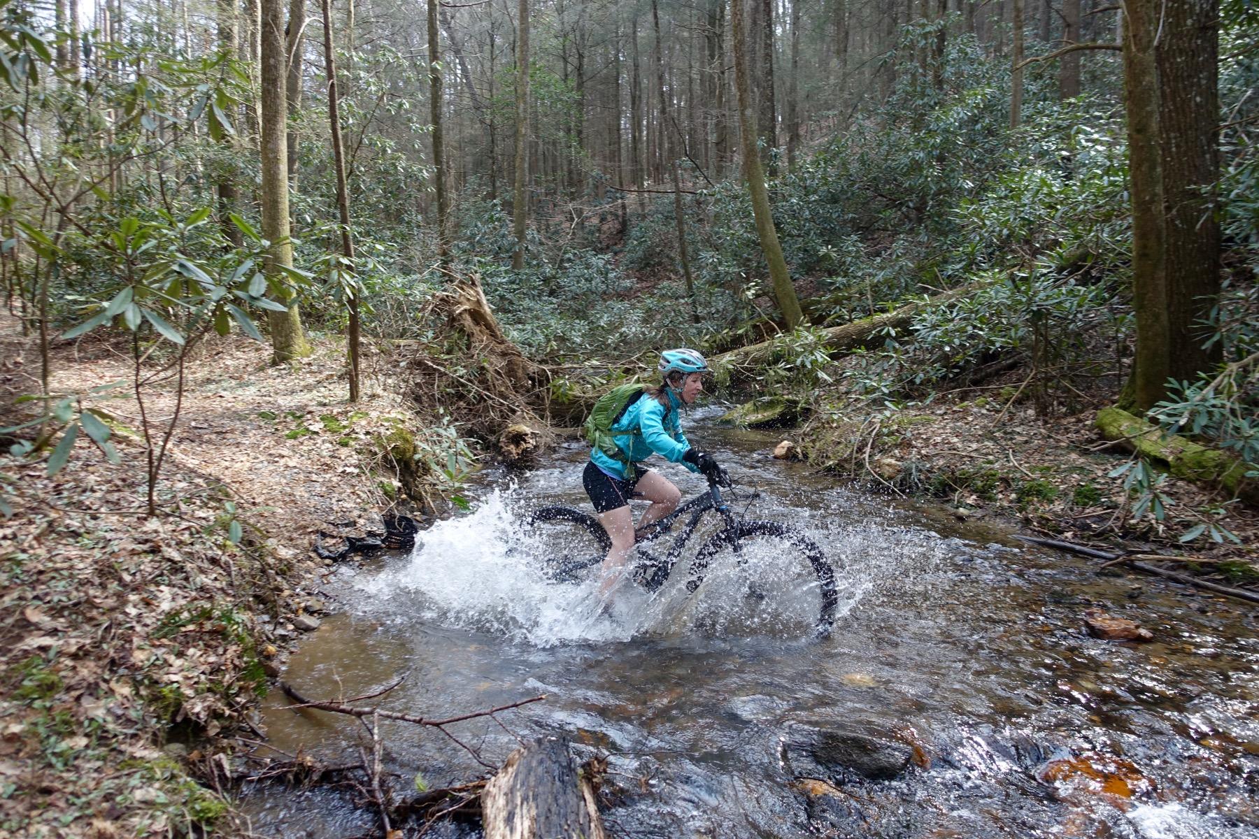 A mountain biker splashes through a shallow stream surrounded by lush green foliage and trees in a forested area. The rider is wearing a helmet and a blue jacket, with a backpack, as they navigate the rocky terrain. Water sprays around the bike, adding a sense of adventure to the scene. Pinhoti Trail: Mountaintown Creek Segment mountain bike trail.