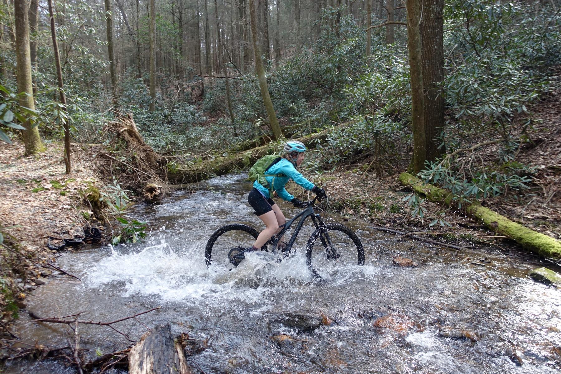 A mountain biker riding through a shallow stream in a wooded area, splashing water as they navigate the rocky path. The rider is wearing a teal jacket, a backpack, and a matching helmet, surrounded by lush green foliage and trees. Pinhoti Trail: Mountaintown Creek Segment mountain bike trail.