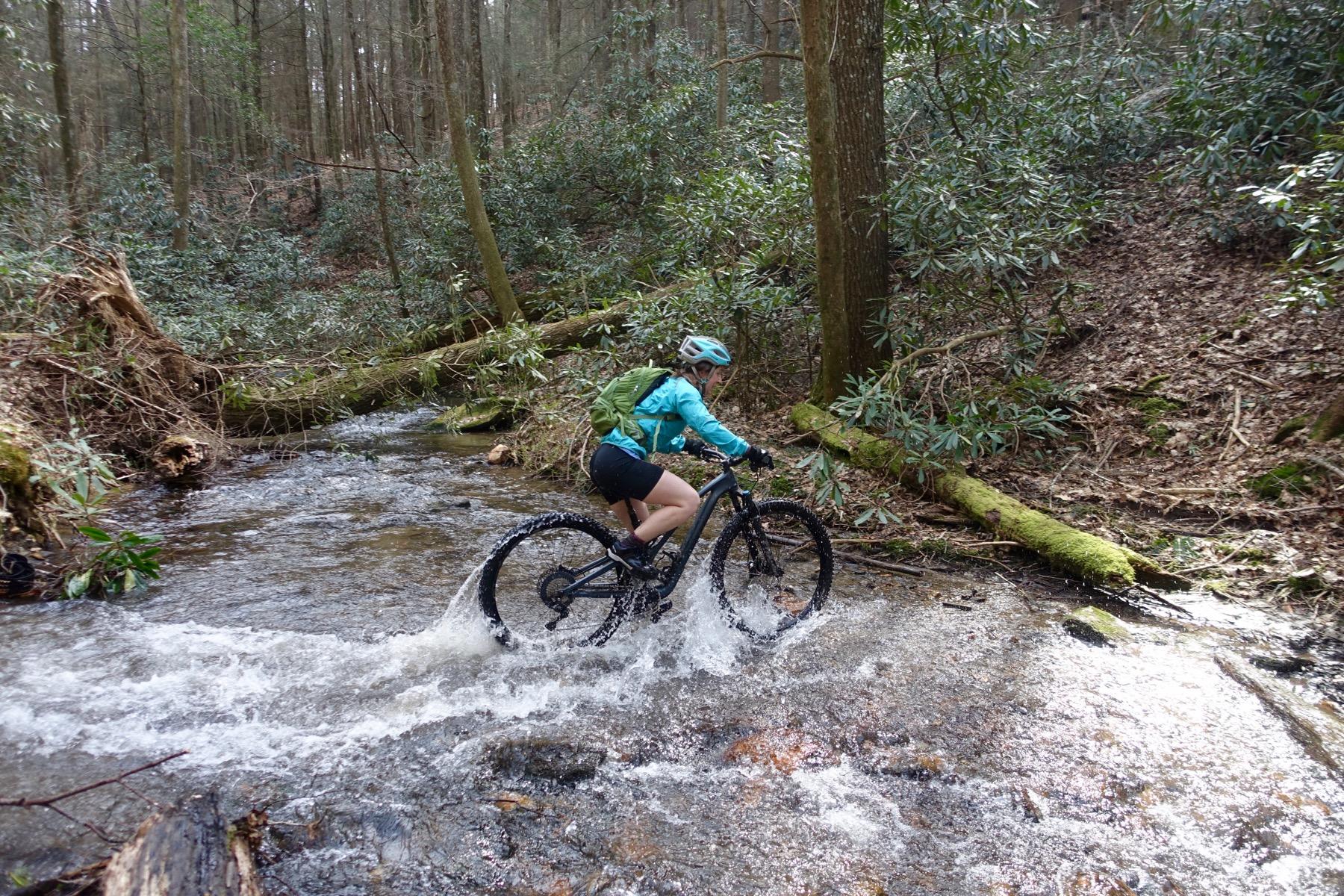 A person riding a mountain bike through a stream in a wooded area, surrounded by trees and greenery. The cyclist, wearing a blue jacket, helmet, and a backpack, splashes water as they navigate over rocky terrain. Pinhoti Trail: Mountaintown Creek Segment mountain bike trail.