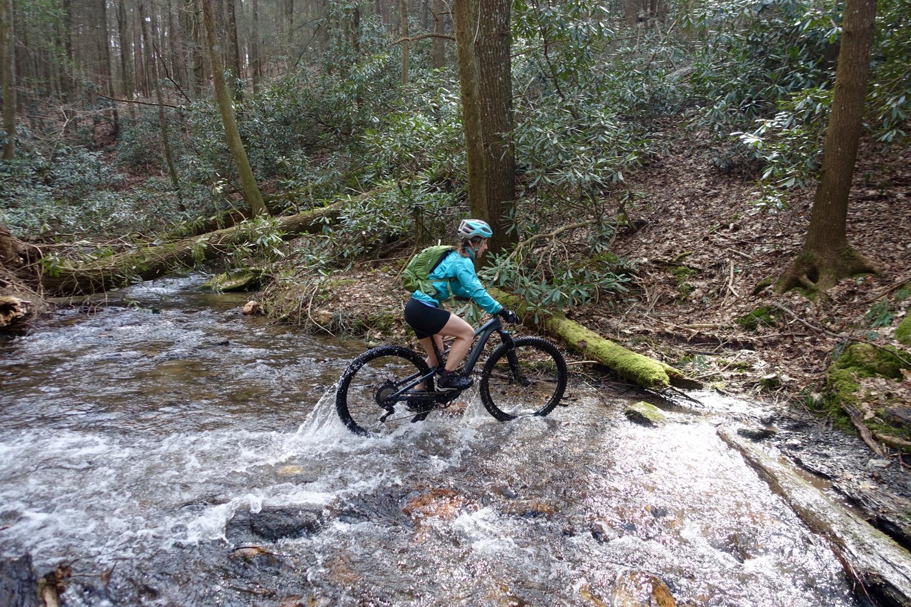 A person riding a mountain bike through a shallow stream in a wooded area, surrounded by trees and greenery. The cyclist is wearing a blue jacket and a helmet, with a backpack secured on their back. Water splashes around the bike tires as it moves through the stream. Pinhoti Trail: Mountaintown Creek Segment mountain bike trail.