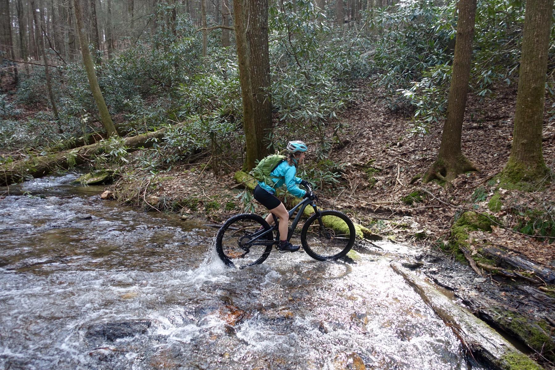 A person riding a mountain bike across a shallow stream in a wooded area, surrounded by lush green foliage and scattered fallen leaves. The cyclist is wearing a blue jacket, black shorts, and a helmet, while a backpack is secured on their back. Sunlight filters through the trees, illuminating the scene. Pinhoti Trail: Mountaintown Creek Segment mountain bike trail.