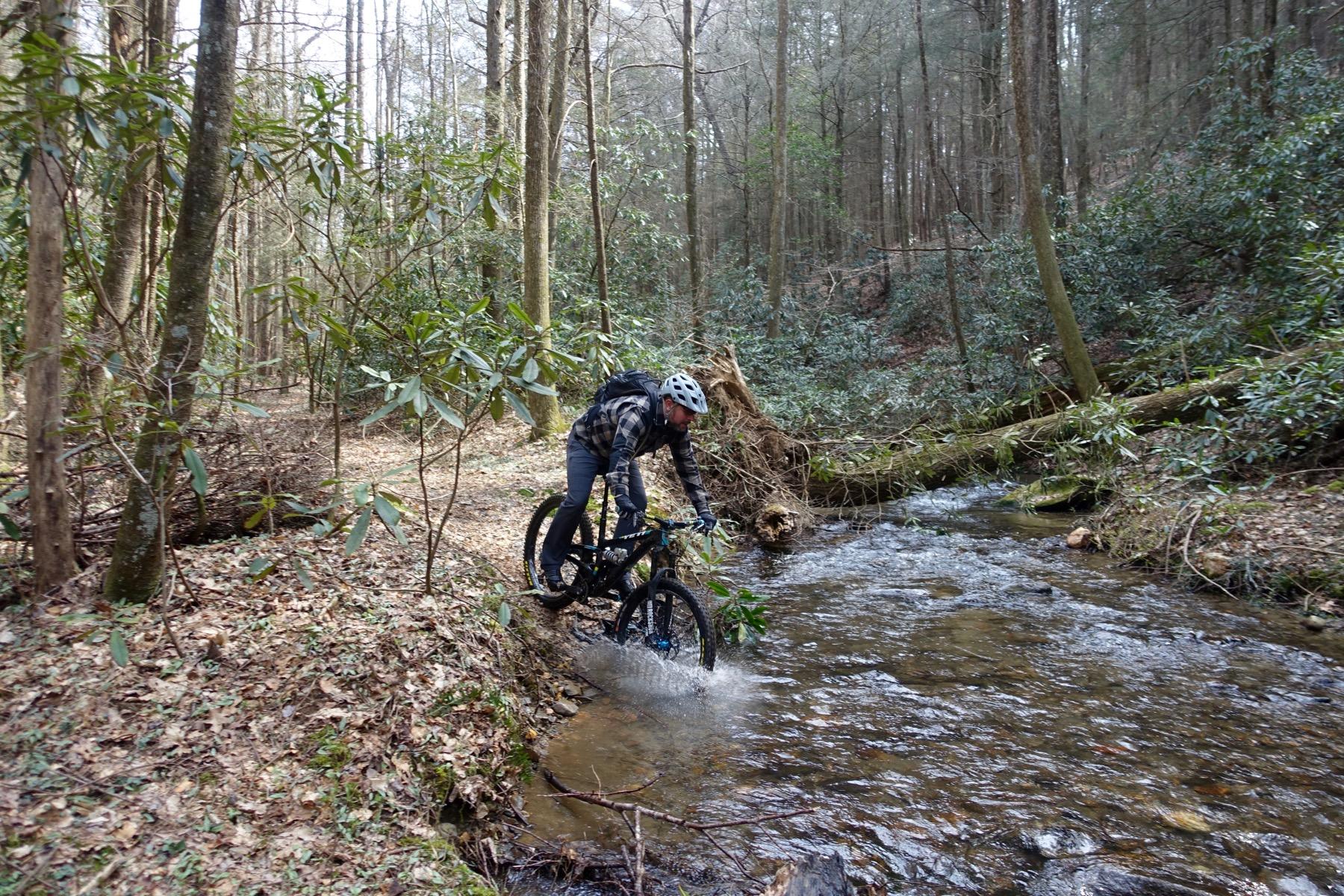 A mountain biker navigating through a creek in a forested area, splashing water as he rides over a rocky edge. Lush greenery and tall trees create a natural backdrop. The biker is wearing a helmet and a plaid jacket, showcasing an adventurous outdoor activity. Pinhoti Trail: Mountaintown Creek Segment mountain bike trail.