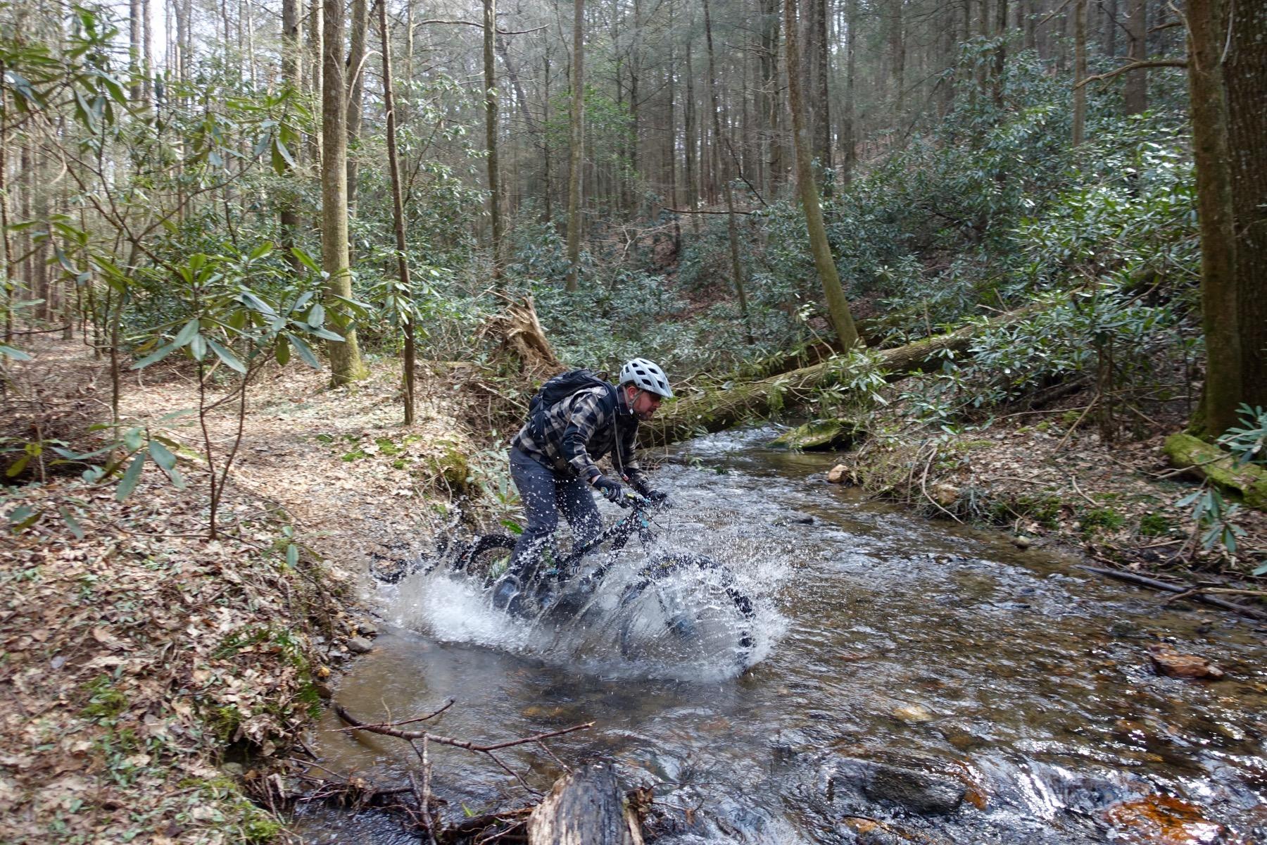 A person riding a mountain bike splashes through a shallow stream in a forest. Surrounding them are tall trees and lush green foliage, with a dirt path visible in the background. Water sprays up from the bike as it navigates the creek, showcasing the excitement of outdoor biking in nature. Pinhoti Trail: Mountaintown Creek Segment mountain bike trail.