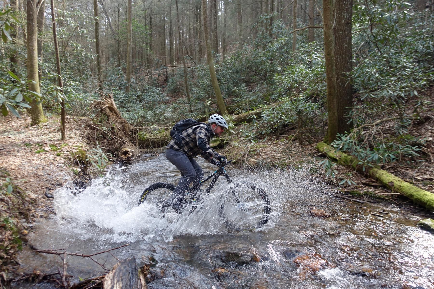 A mountain biker splashes through a stream in a wooded area, surrounded by lush green plants and trees. The rider, wearing a helmet and a plaid jacket, is leaning forward as they navigate over rocks in the water, creating a spray of droplets around them. Pinhoti Trail: Mountaintown Creek Segment mountain bike trail.