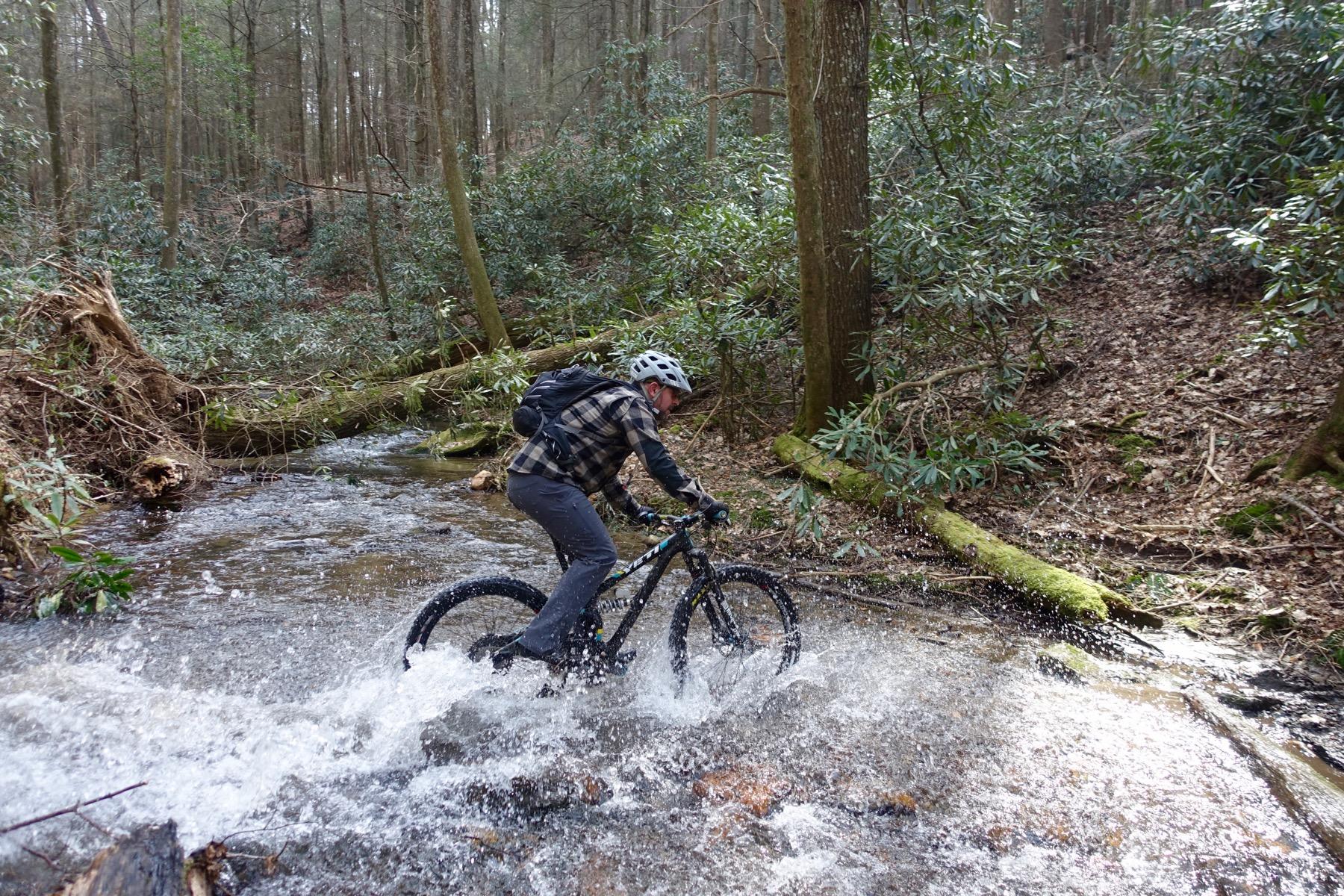 A person cycling through a shallow stream in a wooded area, splashing water as they ride. The cyclist is wearing a helmet and has a backpack, surrounded by trees and greenery. Pinhoti Trail: Mountaintown Creek Segment mountain bike trail.