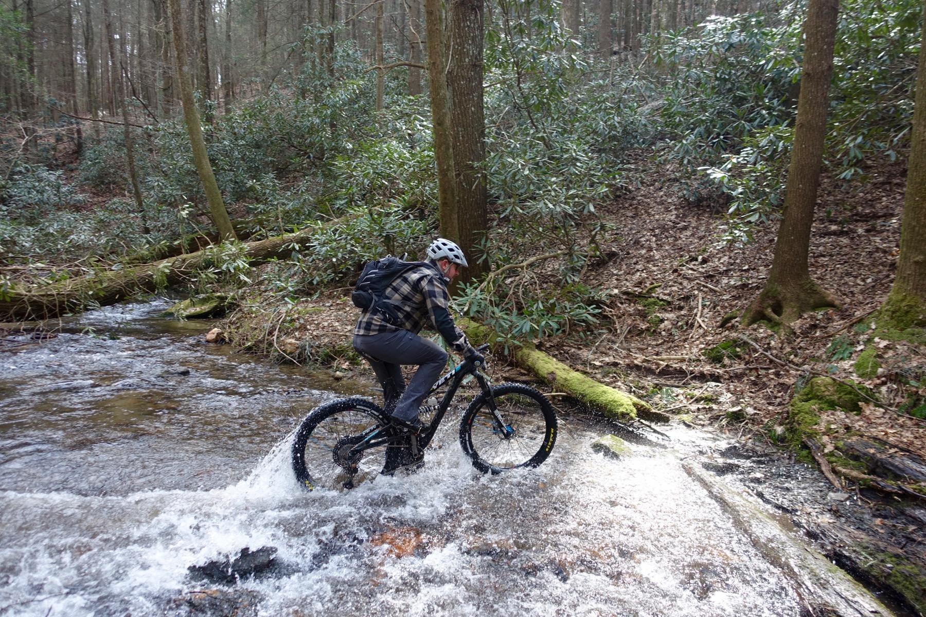 A mountain biker navigates through a shallow stream in a wooded area, with lush greenery and trees surrounding the path. The bike splashes water as the rider maintains balance on the rocky terrain. Pinhoti Trail: Mountaintown Creek Segment mountain bike trail.