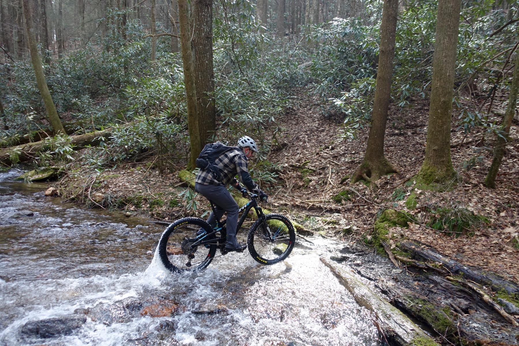 A person riding a mountain bike through a shallow stream in a lush, wooded area, surrounded by trees and greenery. The cyclist is wearing a helmet and a plaid jacket, navigating over rocks and water. Pinhoti Trail: Mountaintown Creek Segment mountain bike trail.