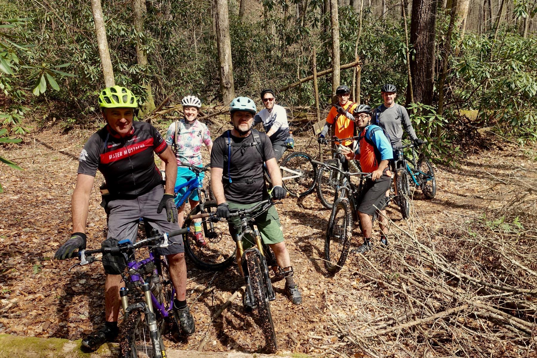 A group of seven mountain bikers, wearing helmets and cycling gear, smile and pose for a photo on a wooded trail surrounded by trees and undergrowth. The cyclists stand next to their bikes, showcasing a variety of colorful clothing and gear, in a sunny outdoor setting. Pinhoti Trail: Mountaintown Creek Segment mountain bike trail.