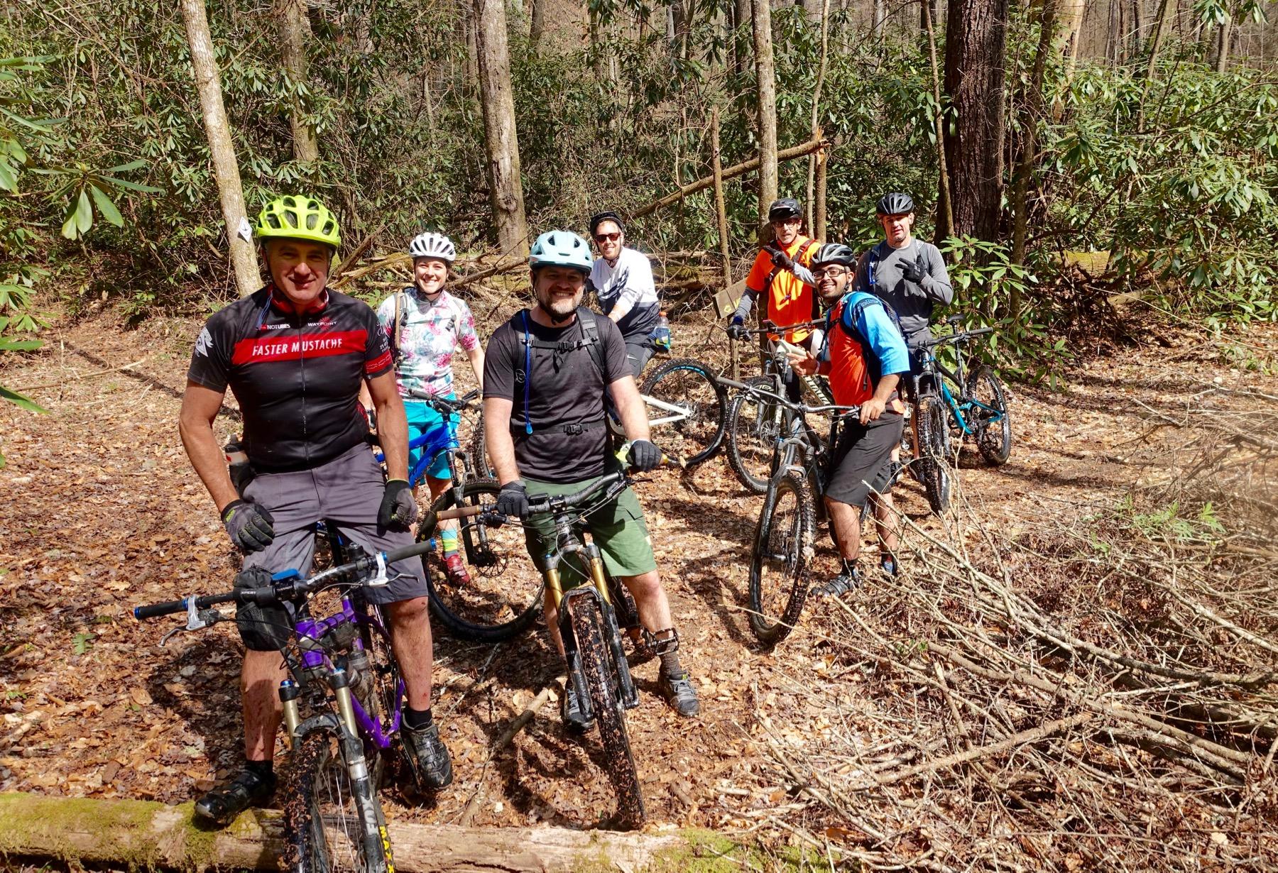 A group of eight mountain bikers posing on a wooded trail surrounded by trees and shrubbery. They are wearing various cycling gear and helmets, with some holding their bikes and others posed beside them. The scene captures a sunny day in a nature setting, with fallen leaves and branches on the ground. Pinhoti Trail: Mountaintown Creek Segment mountain bike trail.