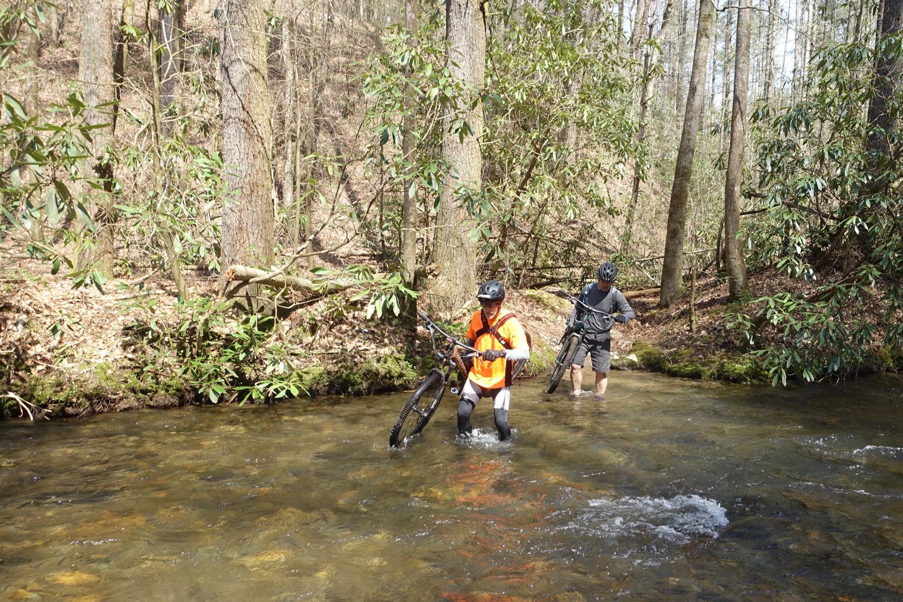 Two mountain bikers crossing a shallow stream in a wooded area. One biker, wearing an orange shirt and carrying their bike, is wading through the water, while the other biker, dressed in a dark outfit, walks alongside them, also carrying their bike. Surrounding them are trees and greenery typical of a forested environment. Pinhoti Trail: Mountaintown Creek Segment mountain bike trail.